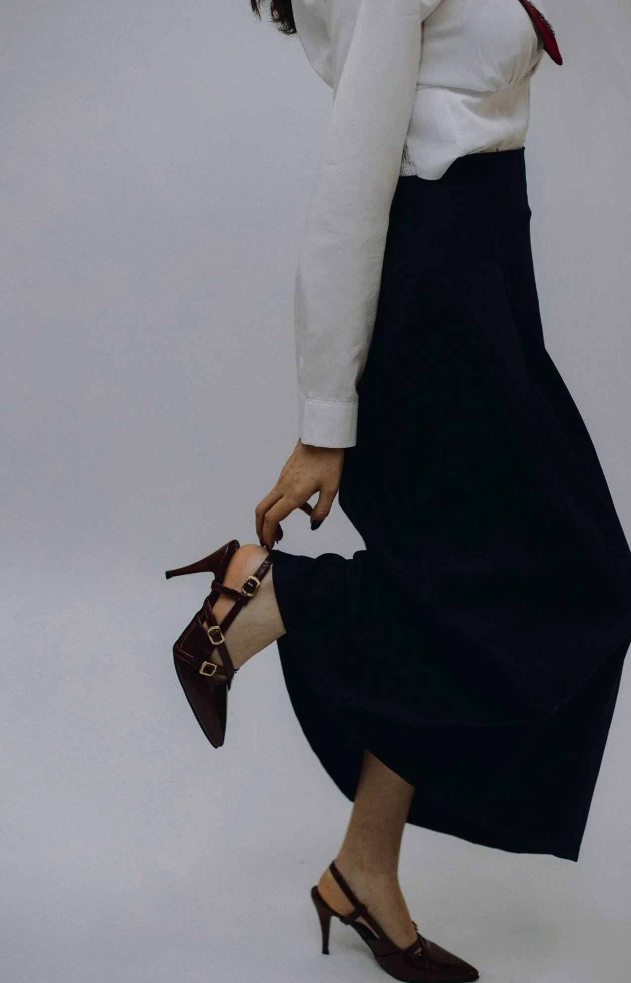 A person dressed in a white blouse, black skirt, and high-heeled shoes, with one leg bent at the knee and foot raised, holding the heel of the shoe with their hand.
