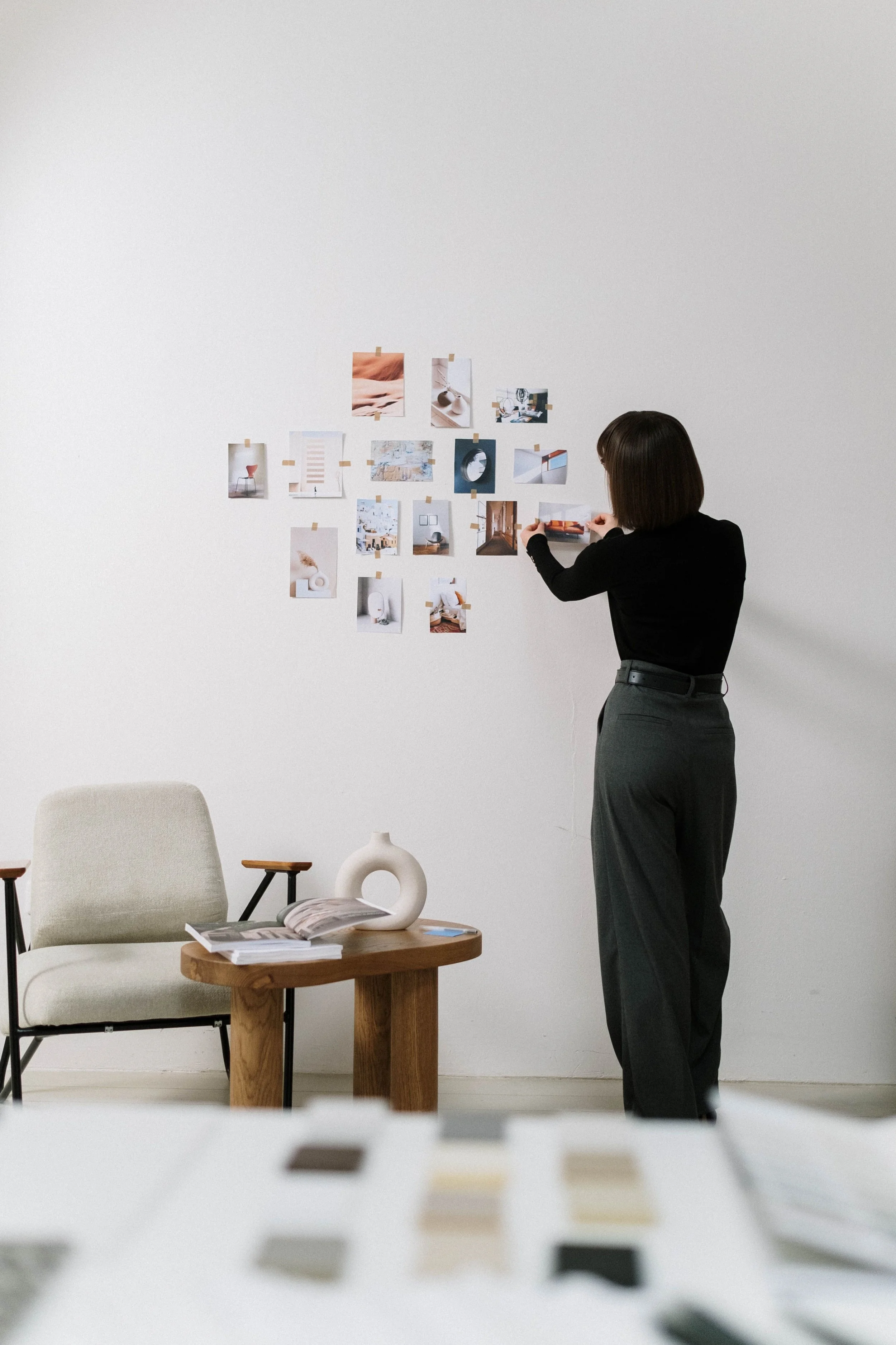 A woman with short dark hair, wearing a black top and gray high-waisted pants, arranging photos on a white wall in a minimalist workspace.