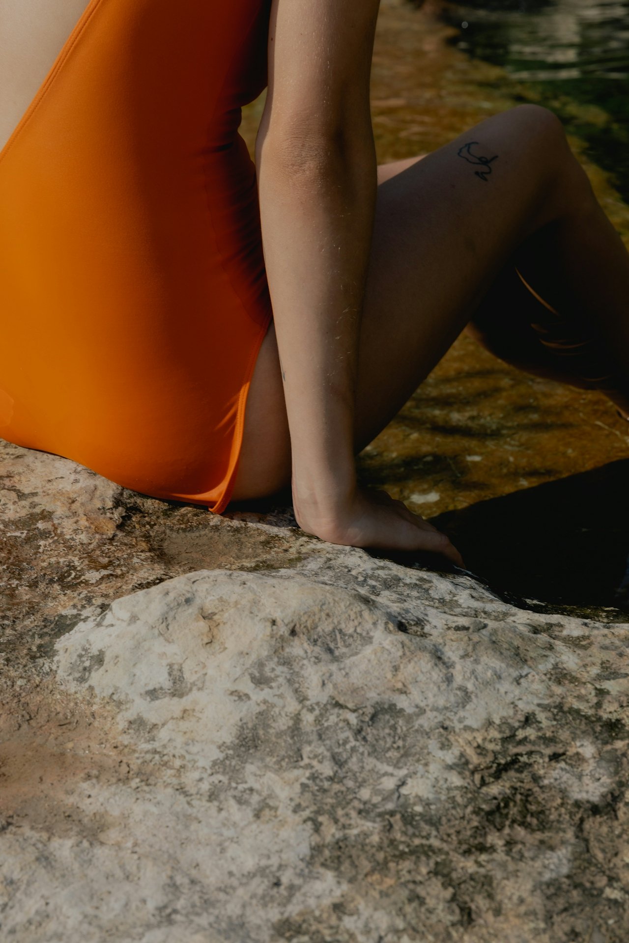 A person wearing orange swimwear sitting on a rock near a body of water, with one hand resting on the rock and the other supporting them. The person has a small tattoo on their thigh and the scene is outdoors, likely by a river or lake.