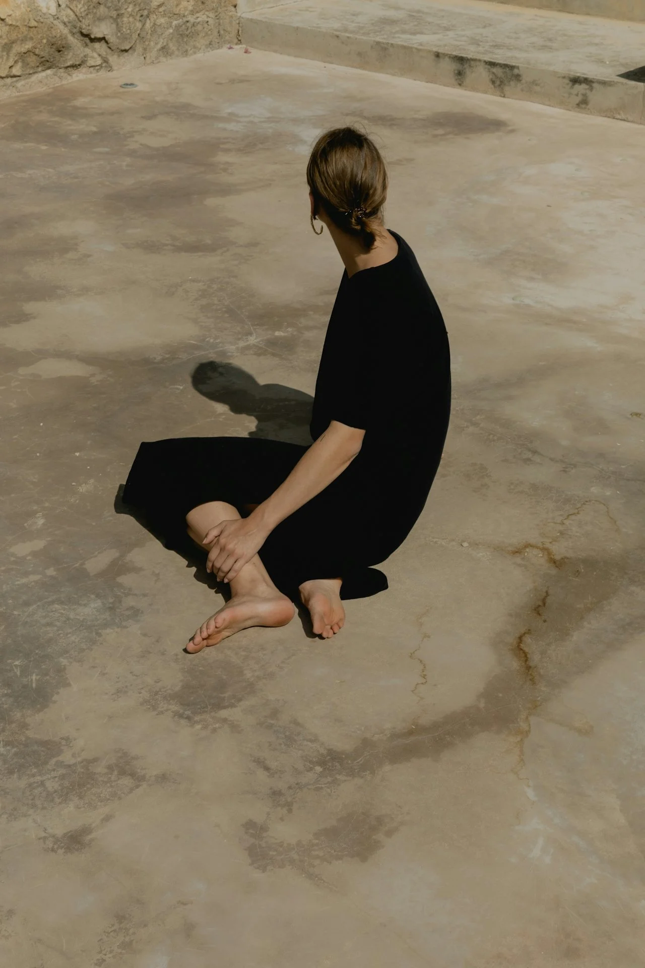 A woman with brown hair in a ponytail, wearing a black dress, is kneeling on a concrete floor with her head turned to the side, casting a shadow on the ground.