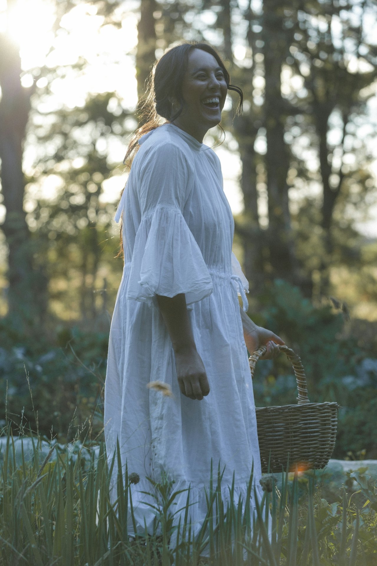 A woman wearing a white dress holding a wicker basket, standing in a forest with sunlight filtering through the trees, smiling and laughing.