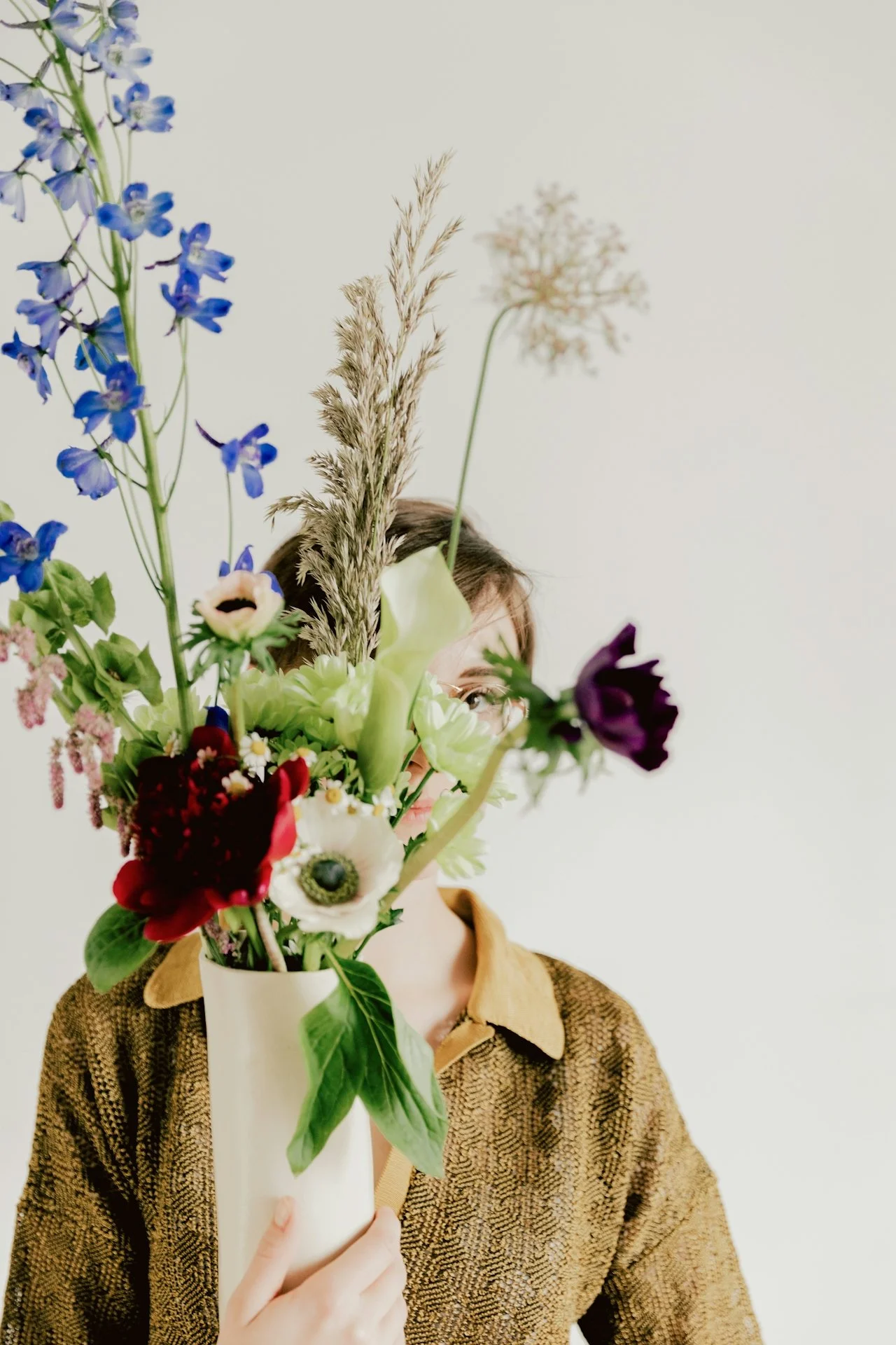 Woman holding a large bouquet of colorful flowers, obscuring her face, against a plain white background.