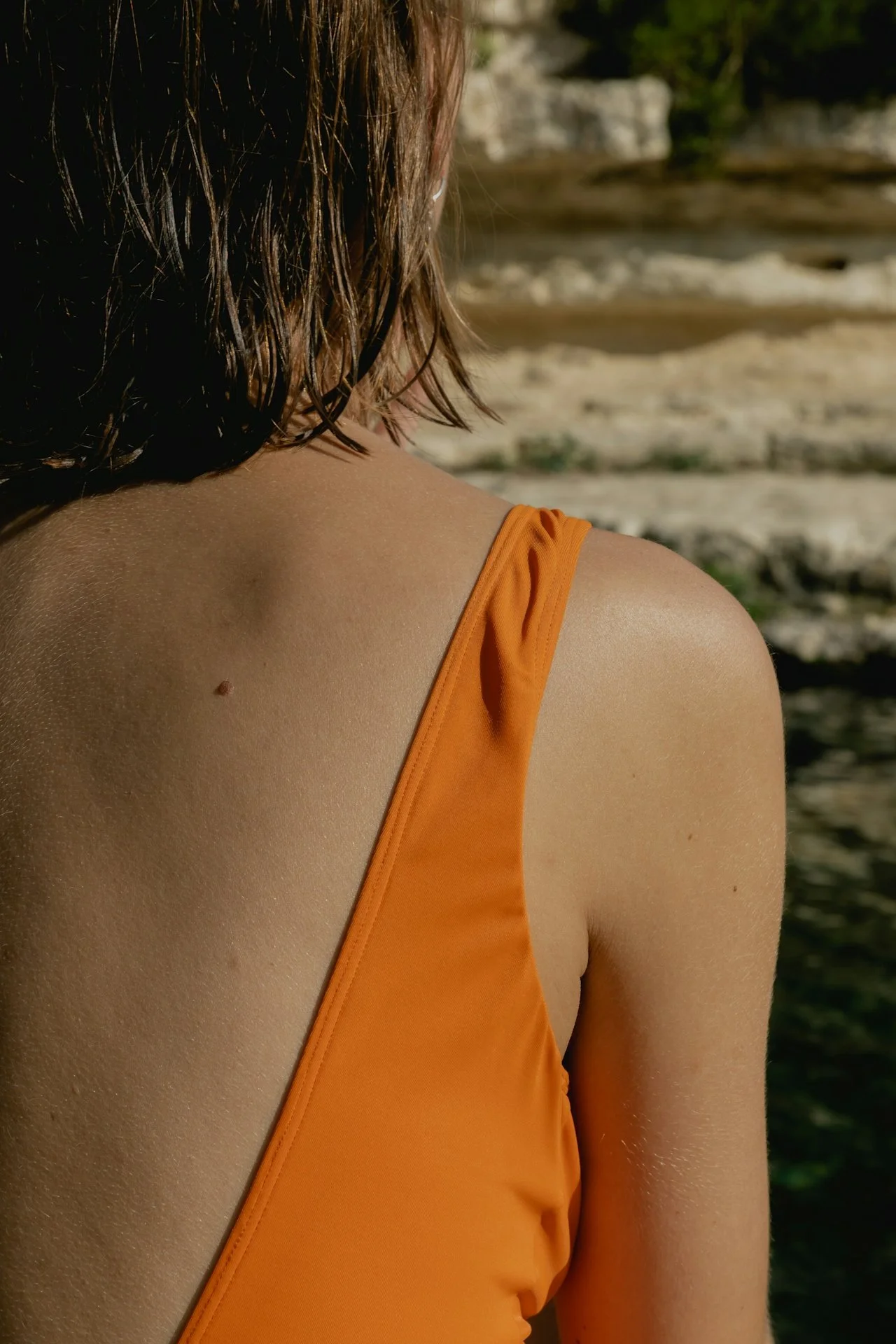 Close-up of a woman's shoulder and back, wearing an orange tank top, with wet, wavy hair and a beach or river background.