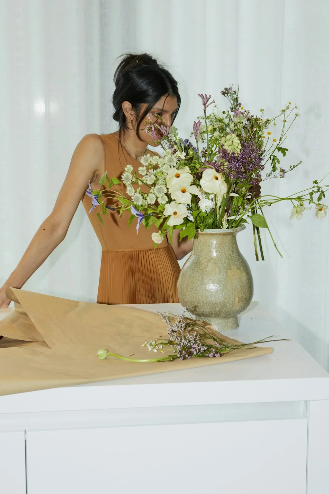 A woman in an orange dress arranges flowers in a large ceramic vase on a white table.