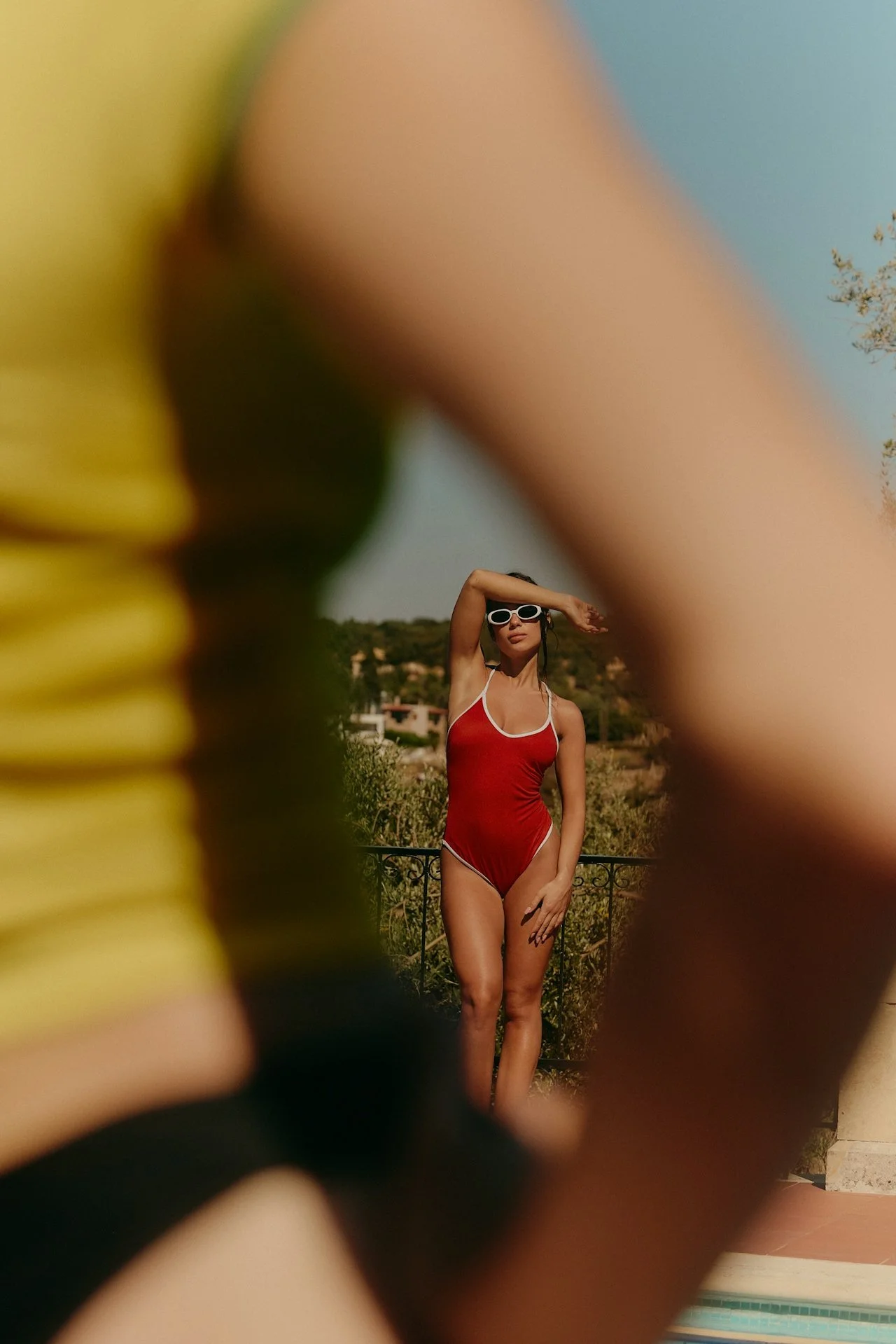 A woman in a red swimsuit and white sunglasses standing outdoors, viewed through a blurred yellow and black object in the foreground.