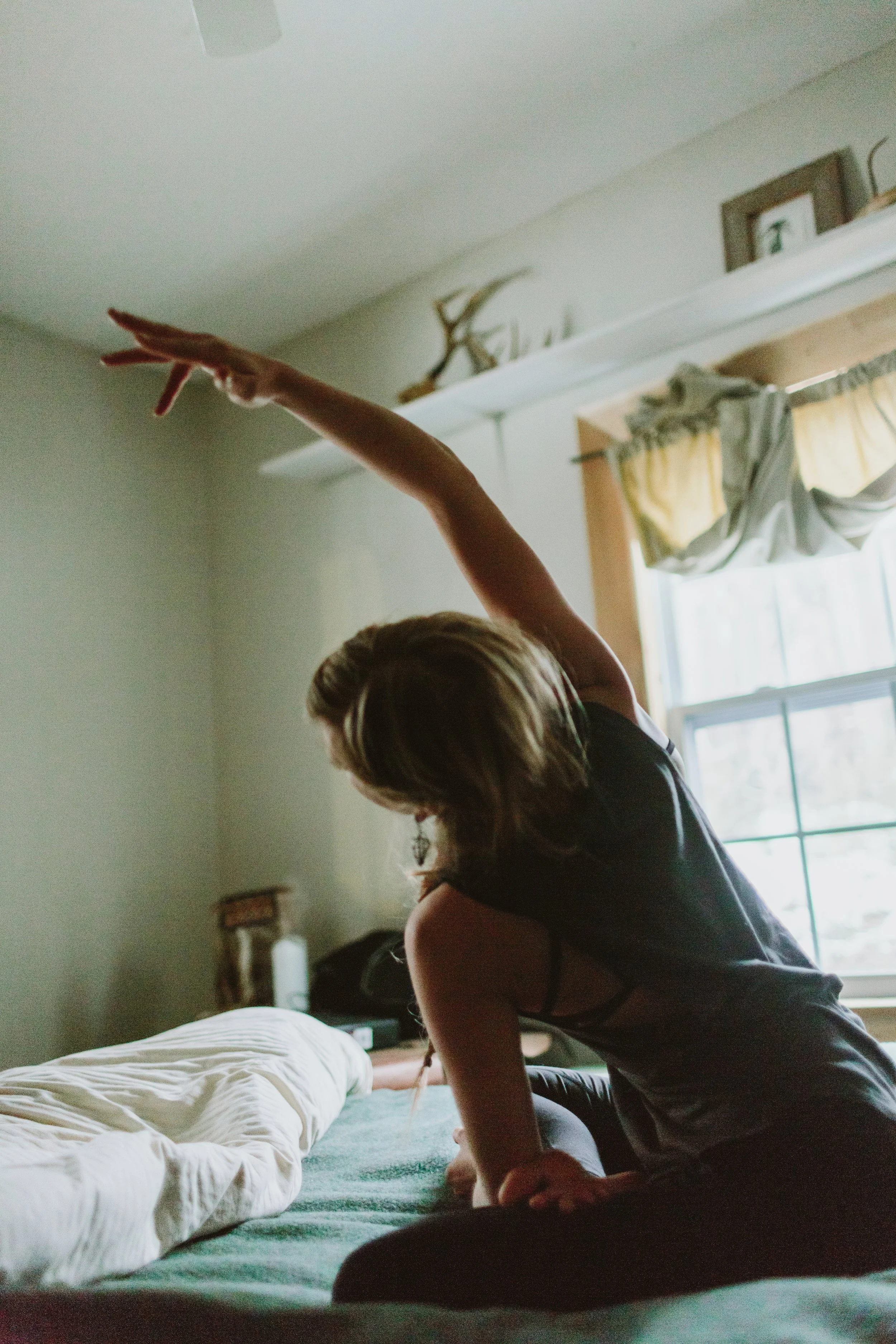 A woman with light brown hair is in a room with natural light, stretching one arm upwards while standing by a bed. The room has a window with curtains, a white shelf with decorative objects, and a white ceiling fan.