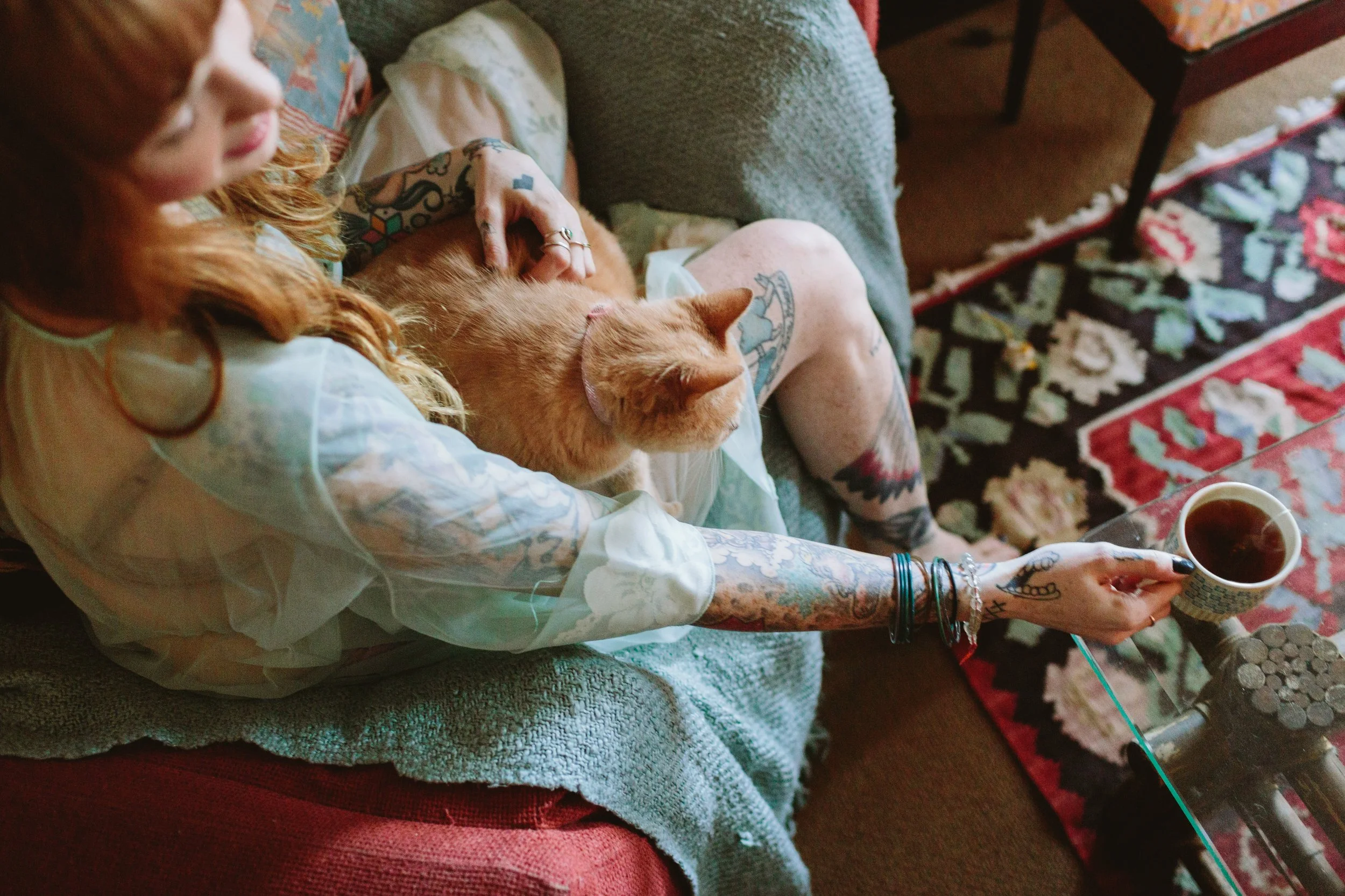 A red-haired woman with tattoos sitting on a couch, holding a mug of tea, with an orange cat resting on her lap and a patterned rug on the floor.