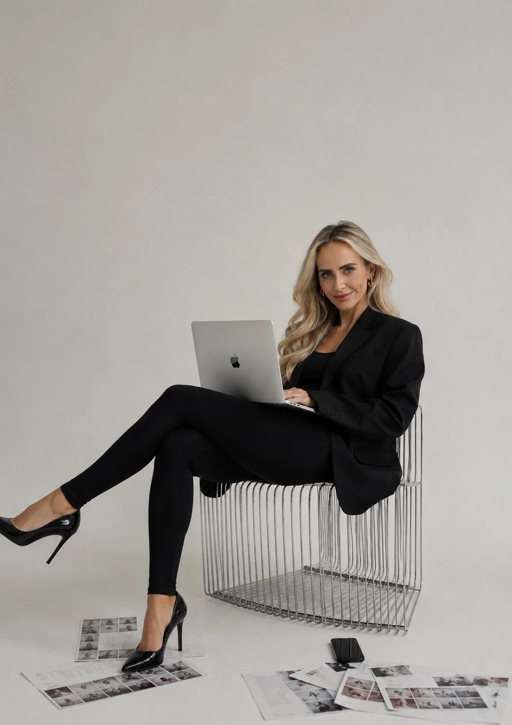 A woman with blonde hair, wearing a black blazer, sitting on a modern wire chair, holding a MacBook laptop, surrounded by scattered papers and a smartphone on the floor, against a plain white background.