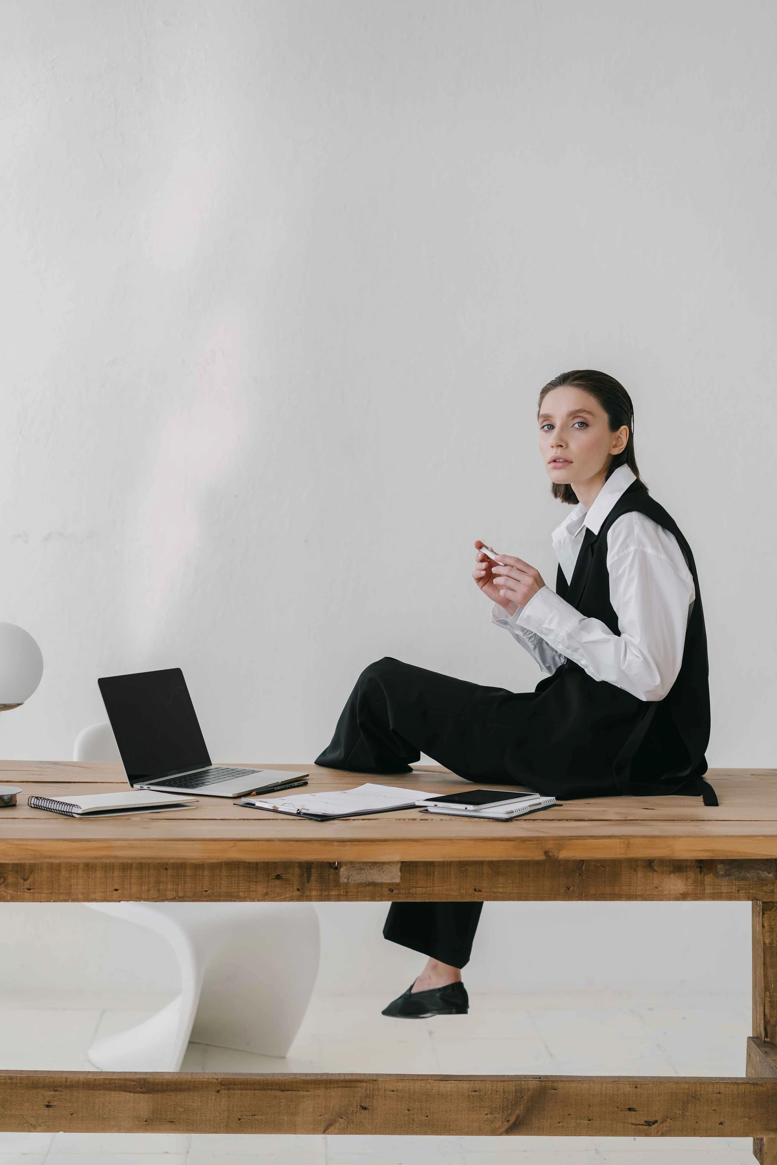 A woman in business attire sitting on a table with her legs crossed, holding a phone, with an open laptop, notebooks, and papers on the table, and a white chair behind her, against a plain white wall.