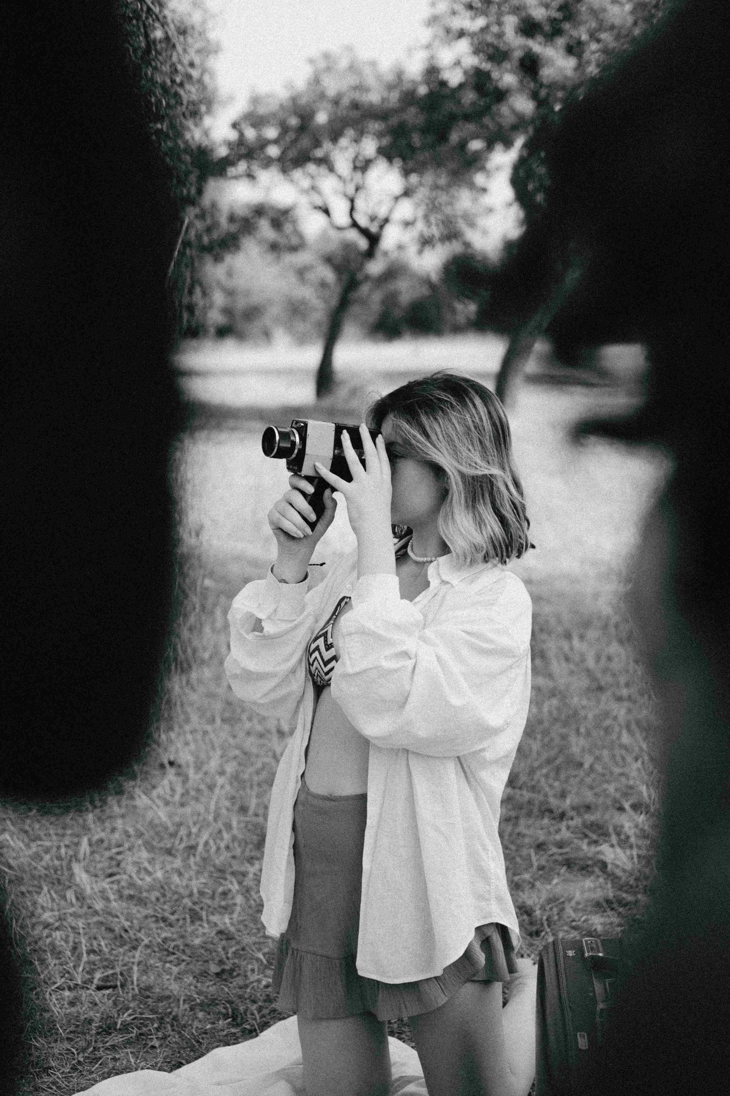 A young woman looking through a camera outdoors in a park, surrounded by trees.