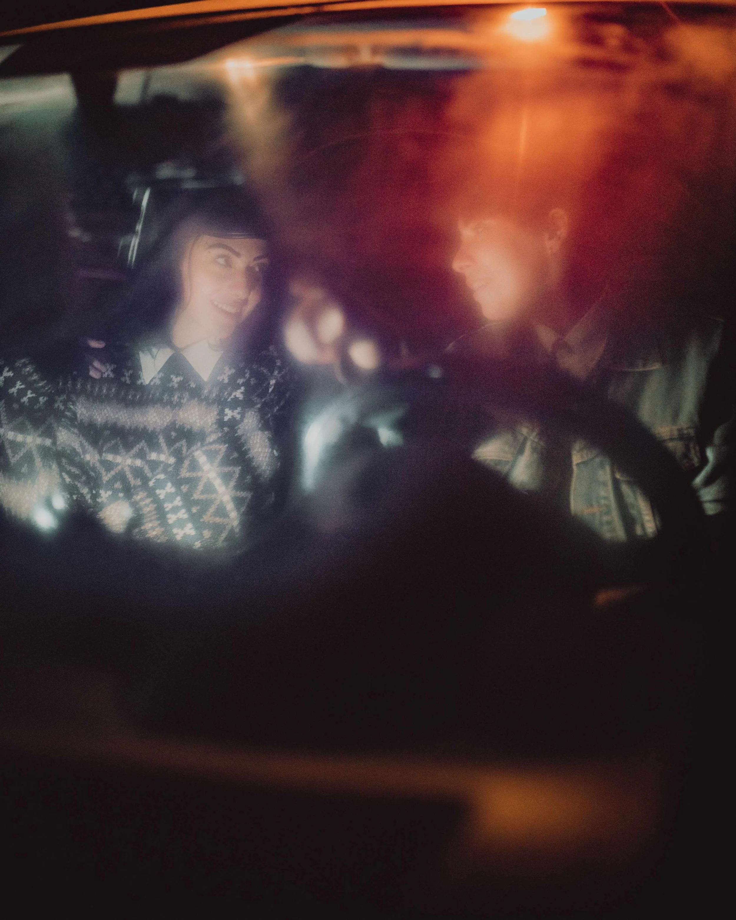 The image shows a view through a car's window at night, with two smiling women inside the car. One woman is pointing towards the camera, and the other is looking at her. The photo has a slight blur and some glare.