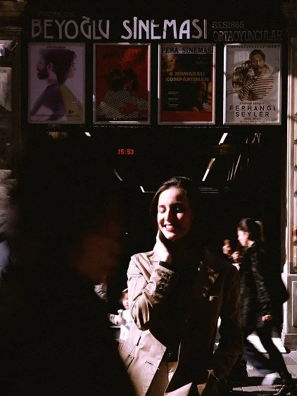 A woman smiling and standing in a crowded area with a theater entrance in the background, displaying posters and a clock showing 15:53.