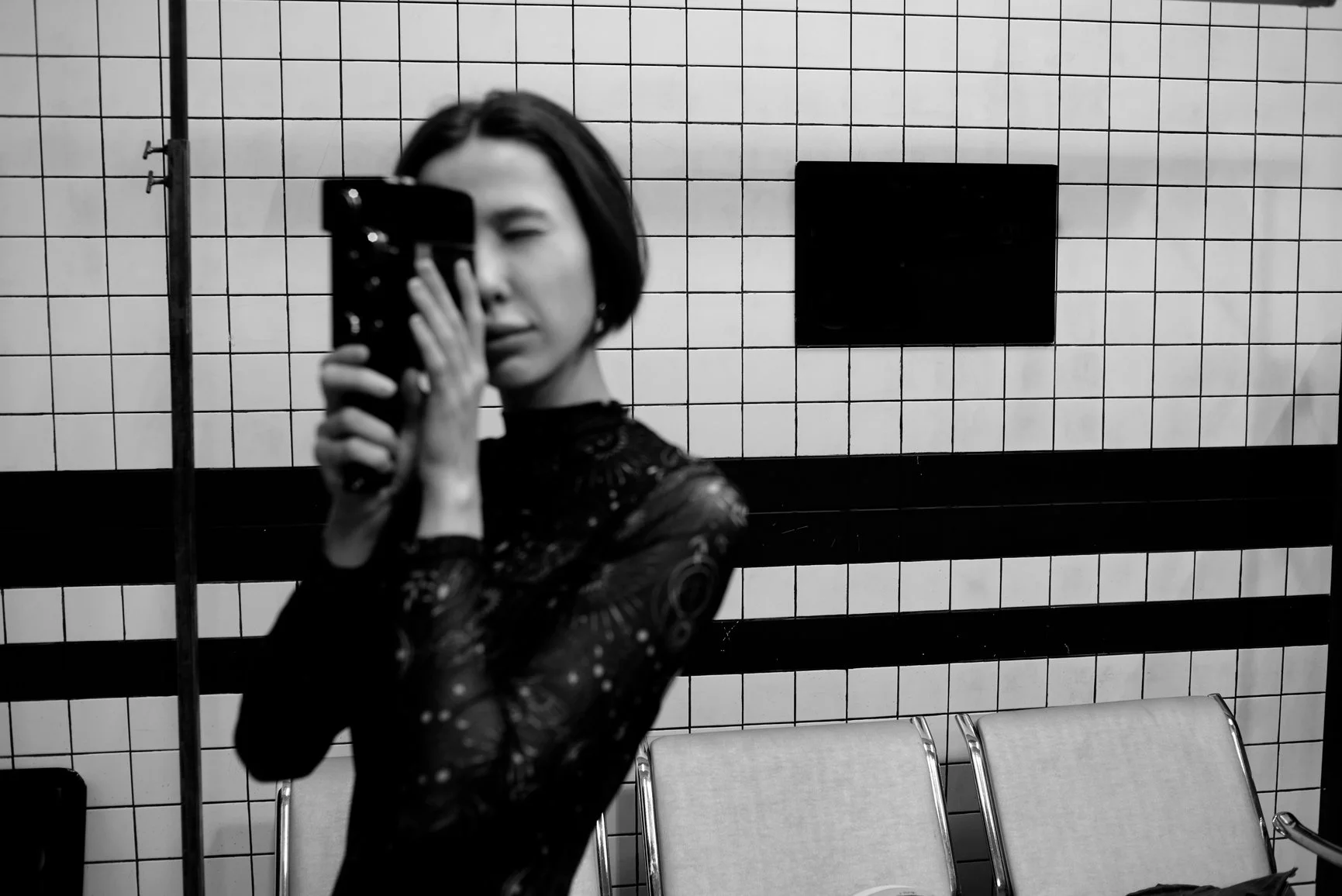 A woman in black lace clothing taking a mirror selfie in a tiled room, with chairs in the background.