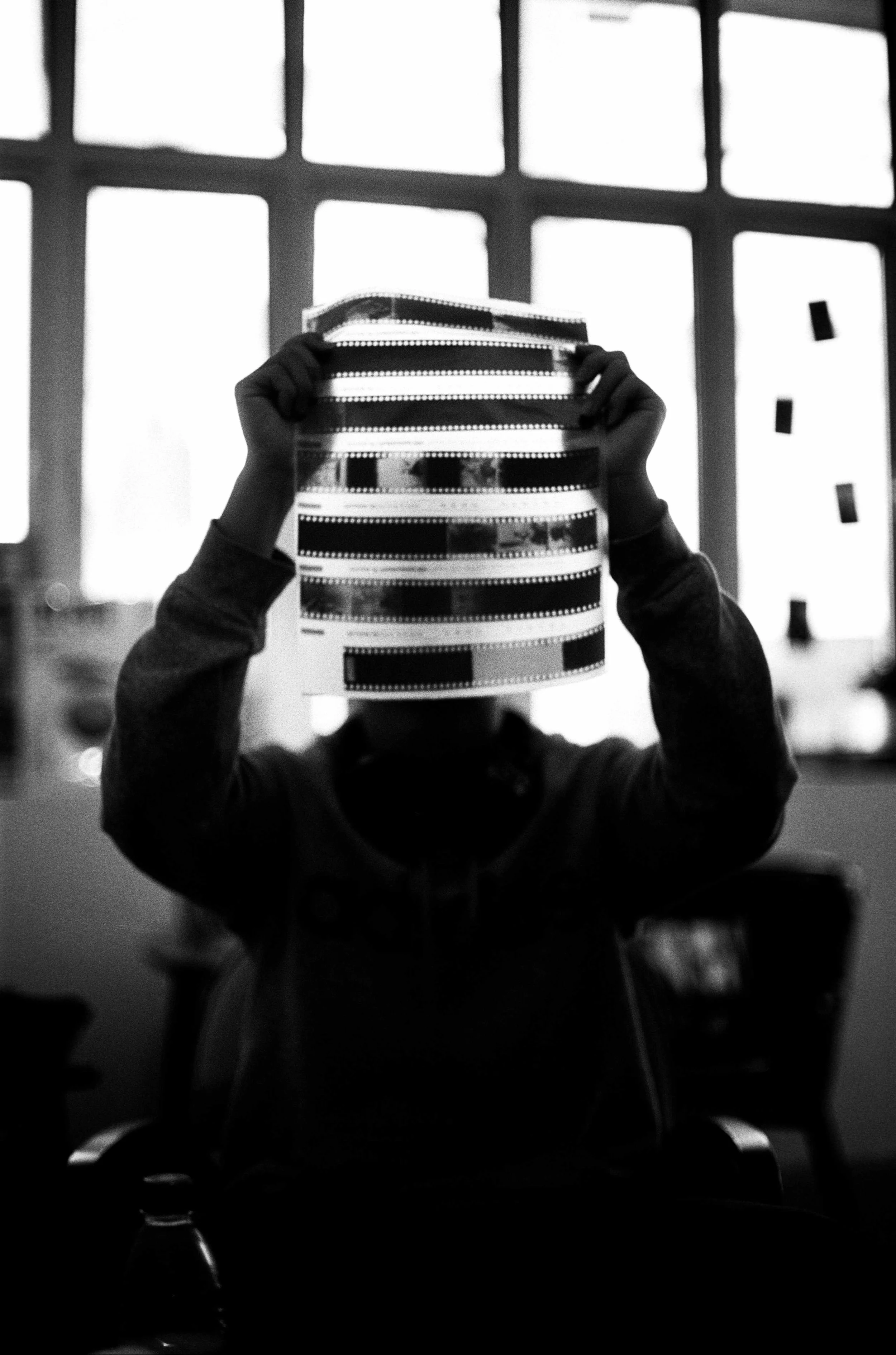 Person holding multiple film negatives in front of a large window, with silhouettes of other objects and a water bottle on a table in the foreground.