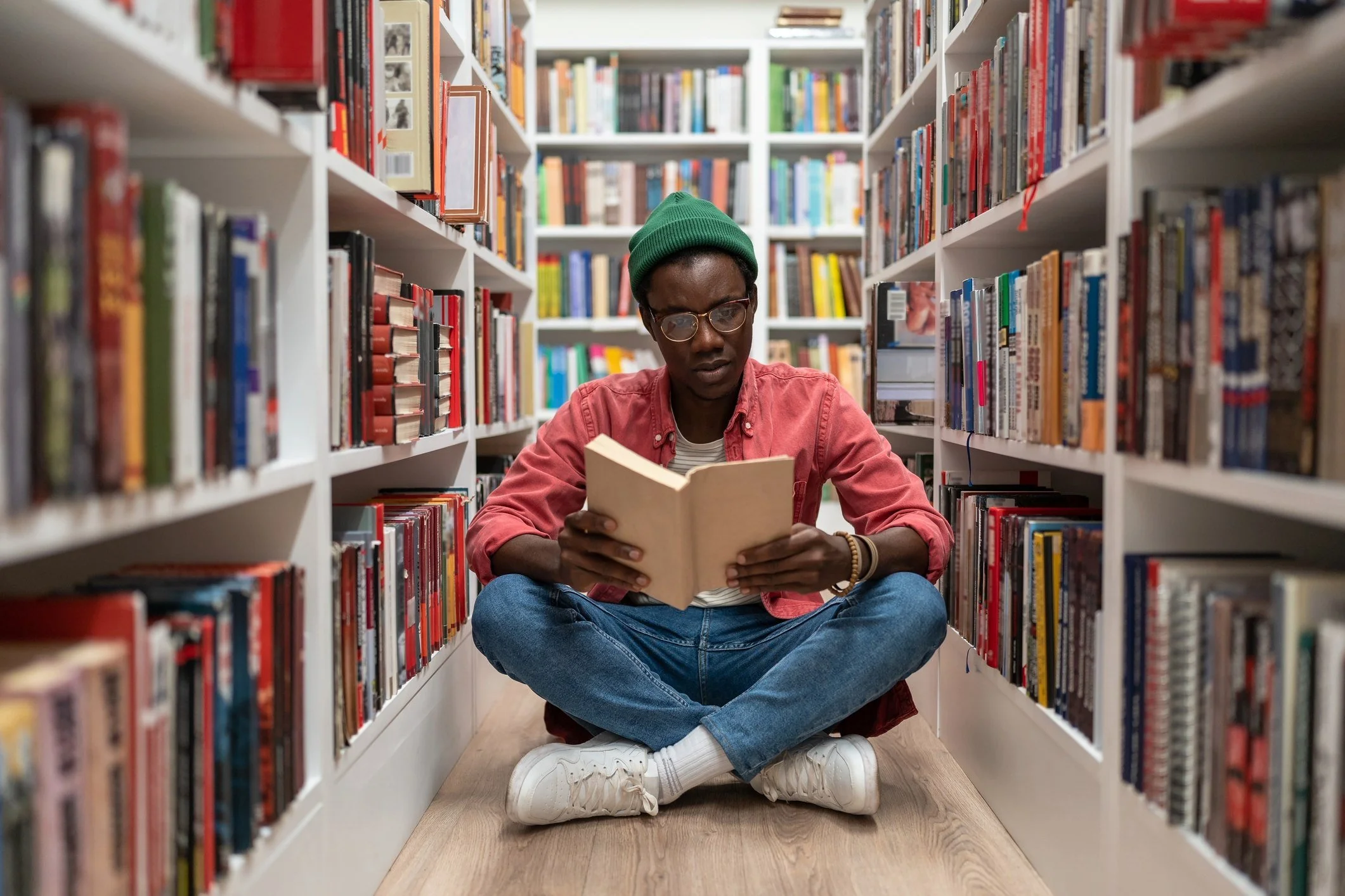 A person sitting cross-legged on a wooden floor in a library aisle, reading a book. They are wearing a red shirt, white sneakers, glasses, a green beanie, and jeans, surrounded by shelves filled with colorful books.