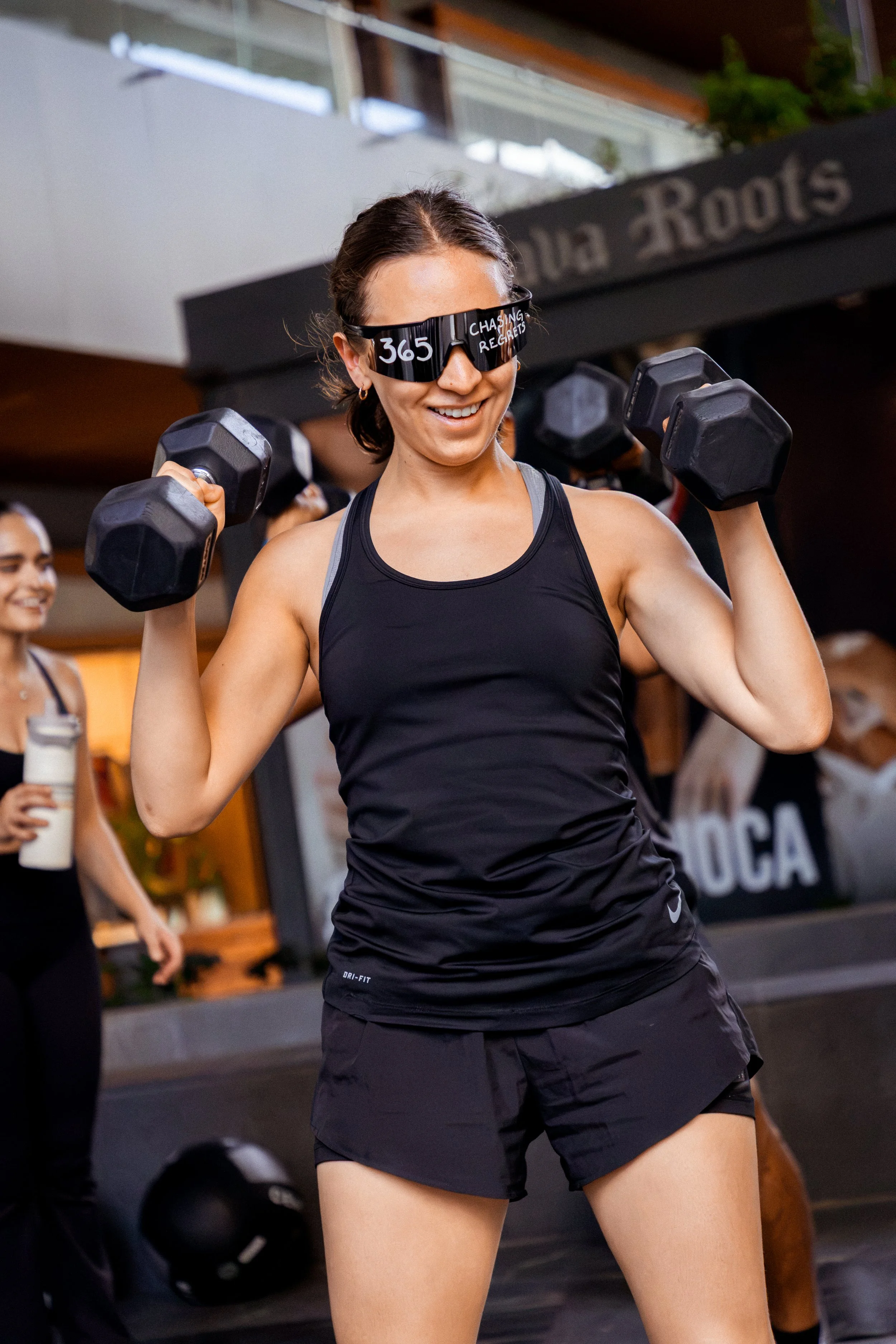 Mujer levantando pesas en el gimnasio, usando gafas de protección con inscripción '365' y la frase 'Chasing Reets', vestida con ropa deportiva negra.