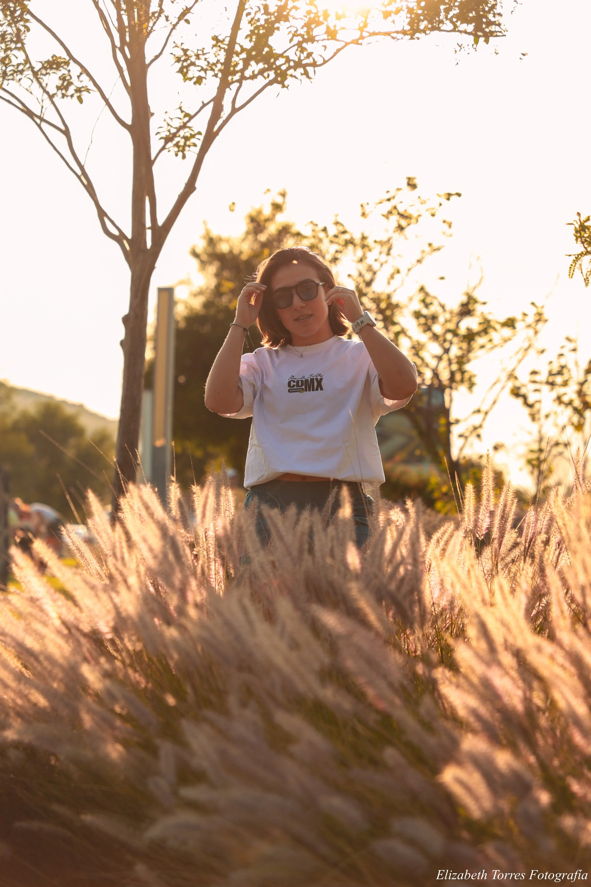 Una mujer con gafas y camiseta blanca con logotipo de CDMX, de cabello castaño, está en un campo de pasto seco durante la puesta del sol, con árboles al fondo y un cielo brillante.