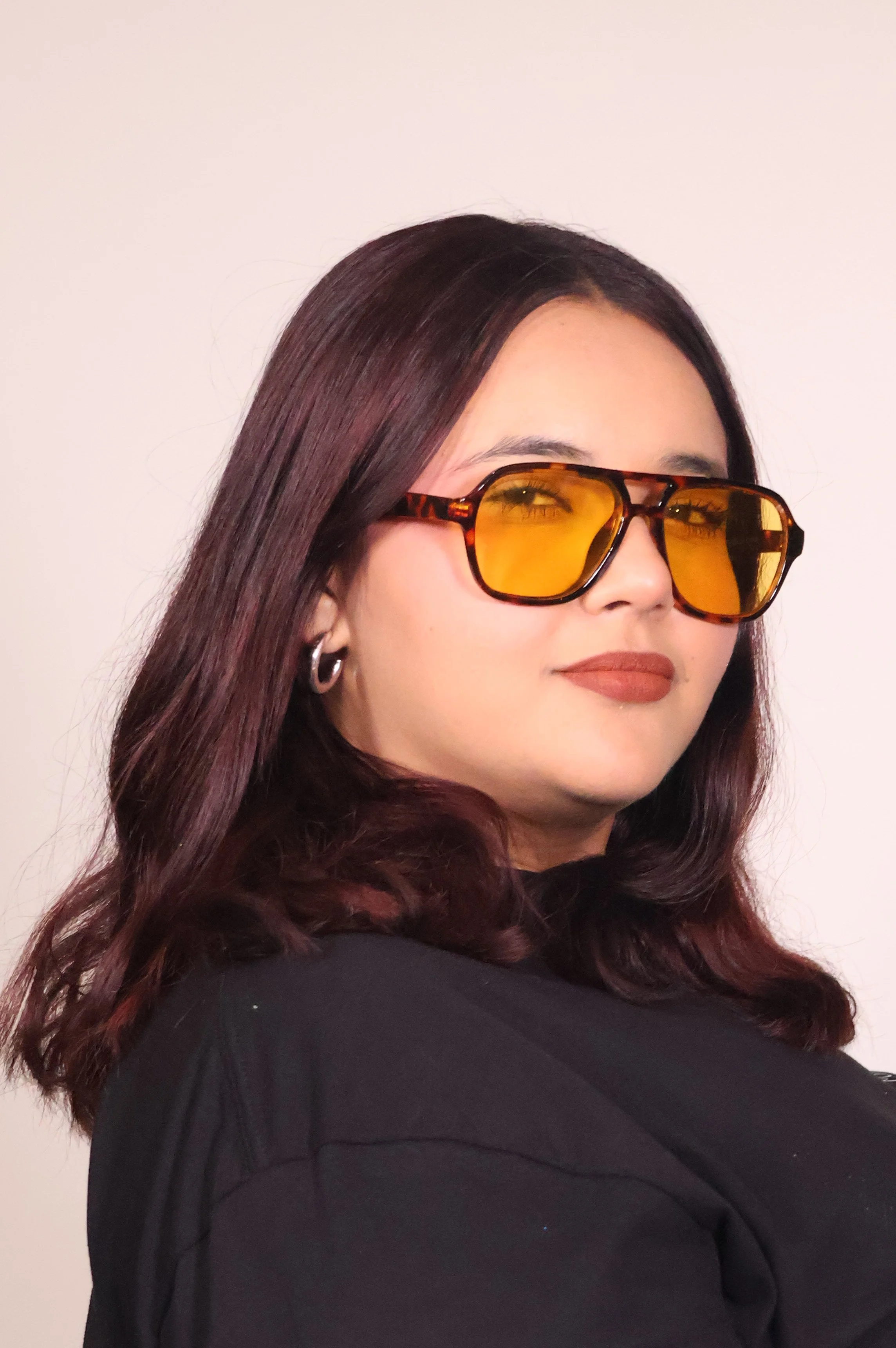 Mujer con cabello oscuro rizado, gafas de sol con lentes amarillos, labios pintados y usando una prenda negra, posa de perfil con fondo blanco
