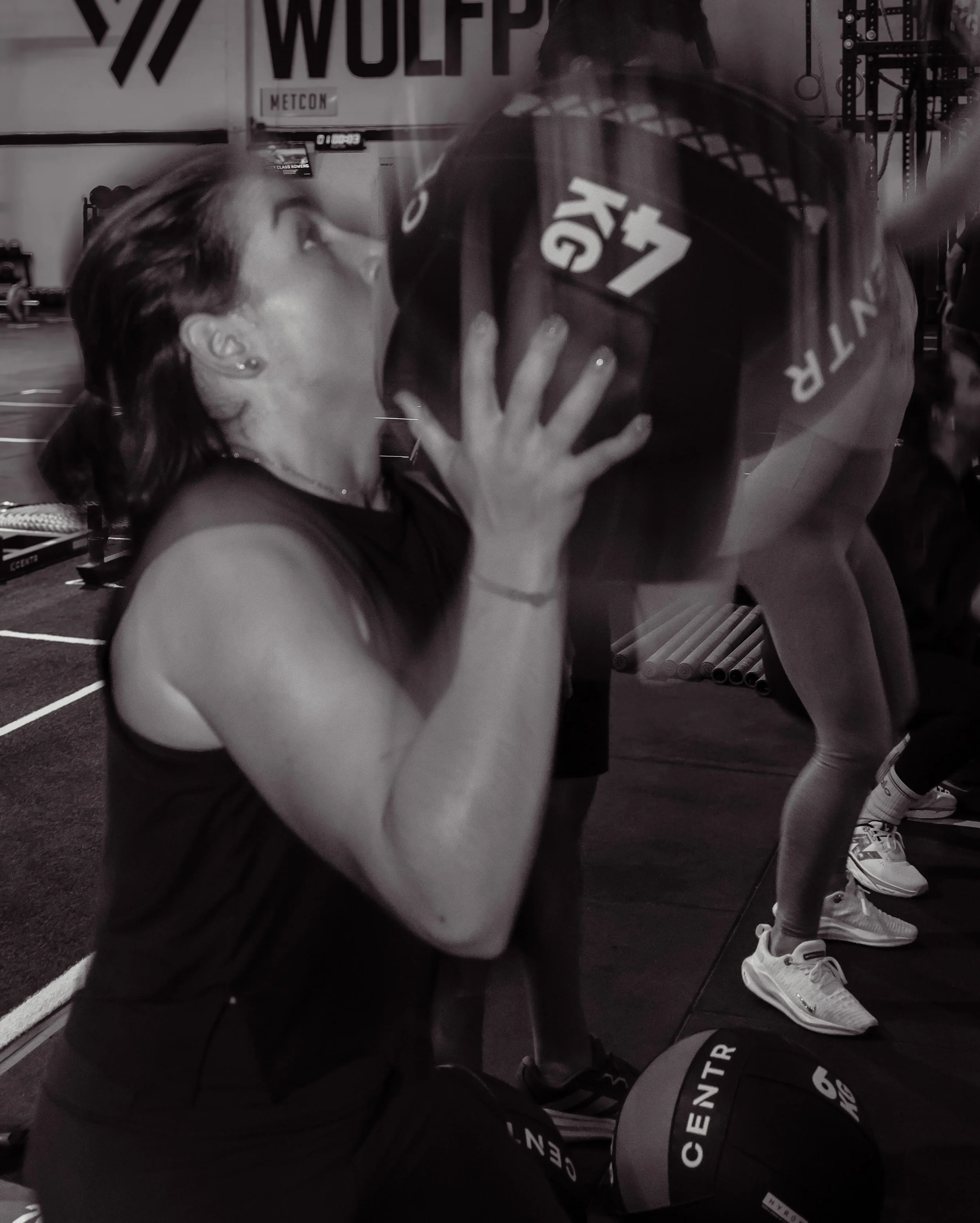 Una mujer en una gimnasia de entrenamiento con una mujer que sostiene una pesa de disco, en un gimnasio con equipo y una pared con logotipos y letras.