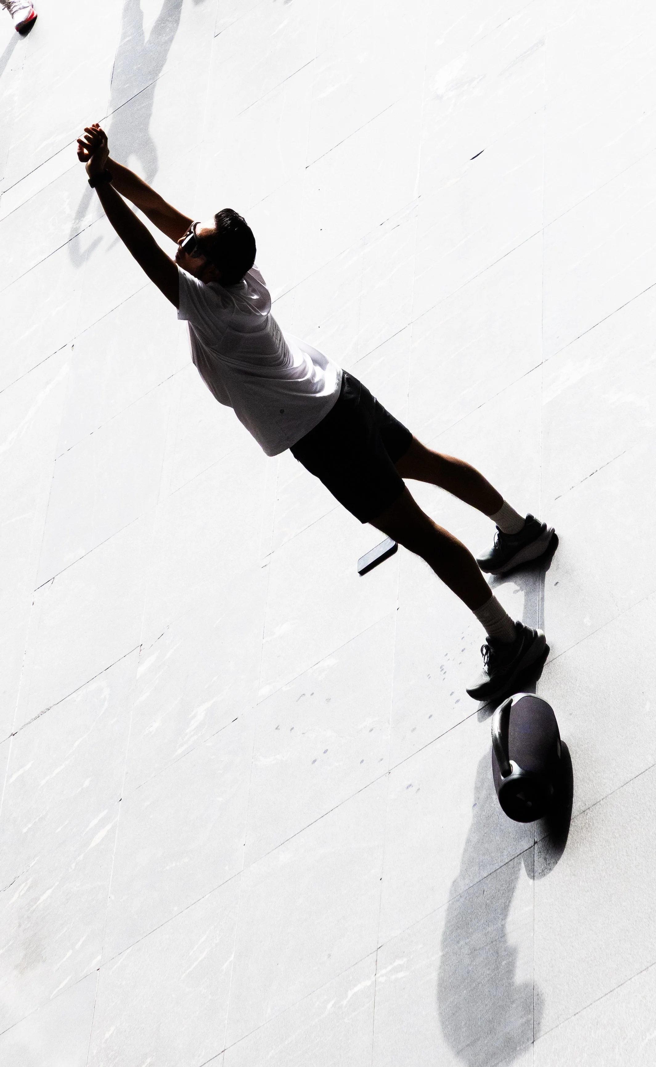 Persona haciendo estiramiento en un suelo brillante, vestido con camiseta blanca, pantalones cortos y gafas, con una kettlebell cerca en sombra.