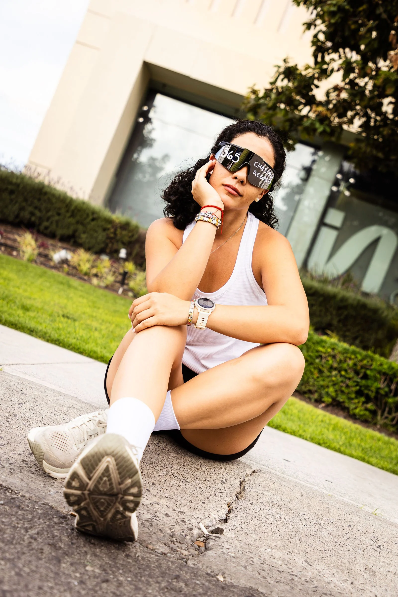 Joven mujer con gafas de invierno, vestido blanco, pantalones cortos negros, zapatillas blancas, y reloj, sentada en la acera con un edificio y arbustos detrás, haciendo una pose casual.