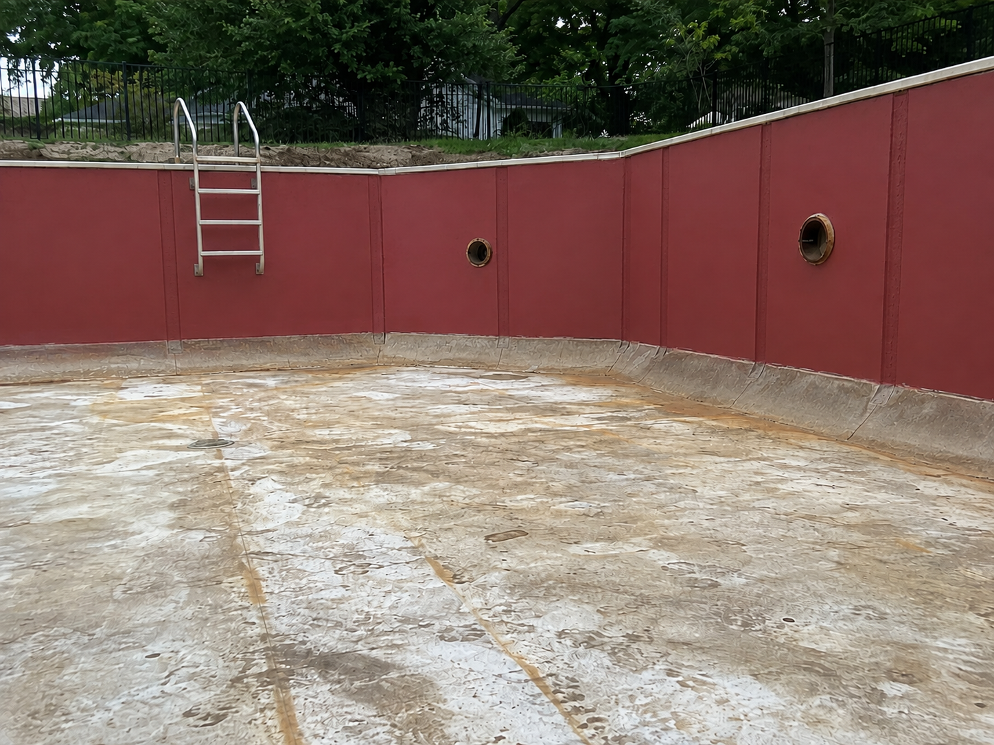 Empty swimming pool with red walls, metal ladder, and two circular openings in the wall, surrounded by trees and black fence.