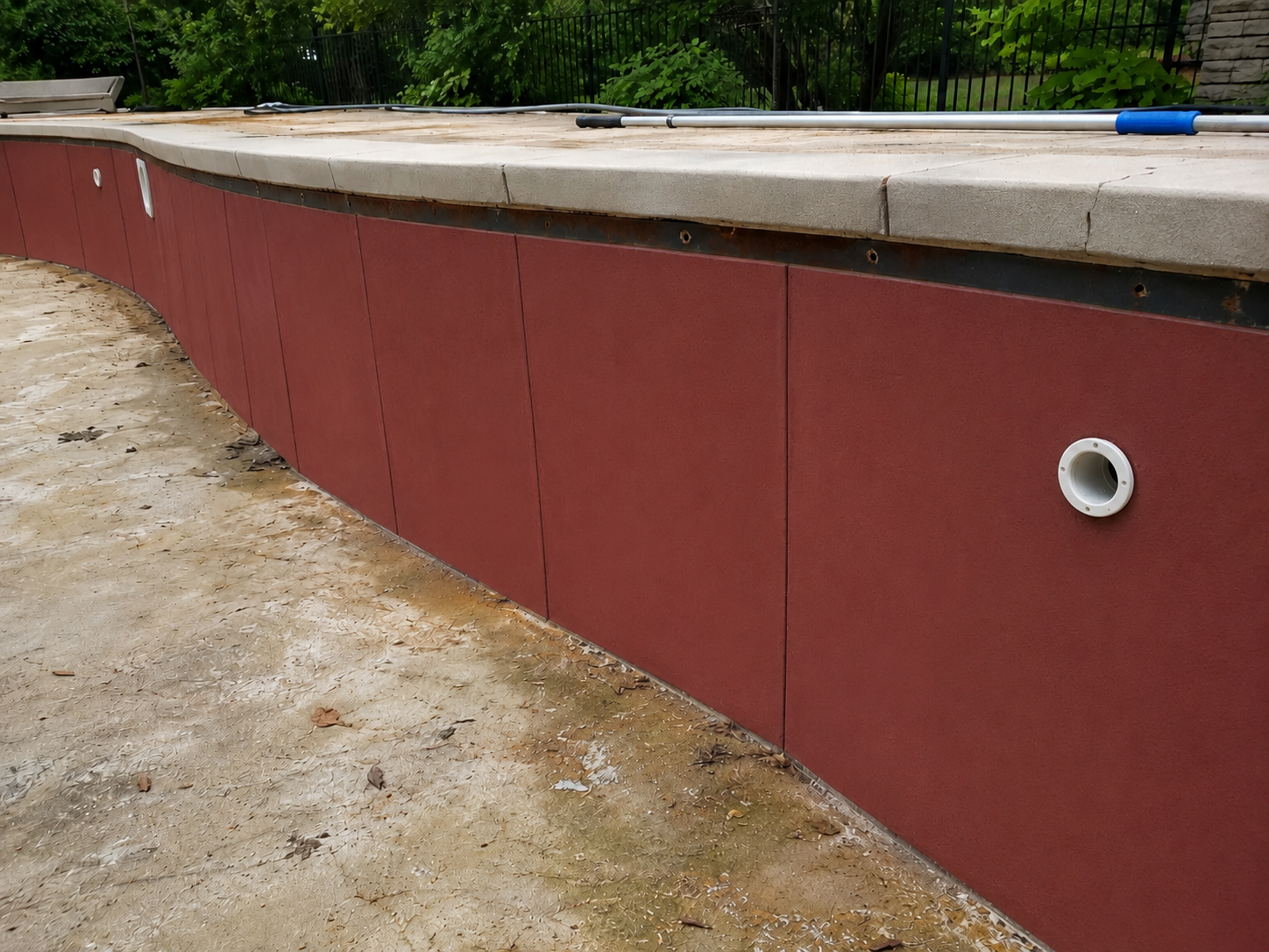 The image shows the side of an outdoor pool under construction with a red board wall, a concrete ledge at the top, and a brown dirt ground with some leaves and debris.