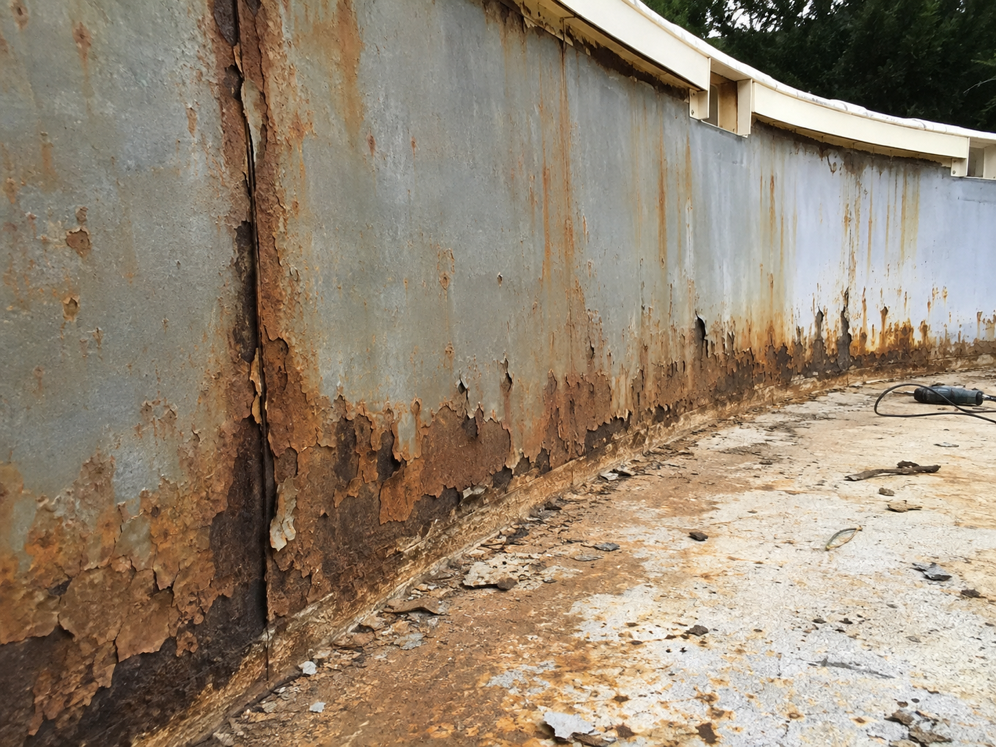 Close-up of a rusted and peeling metal wall with rust stains and corrosion, with power tools and debris on the ground.