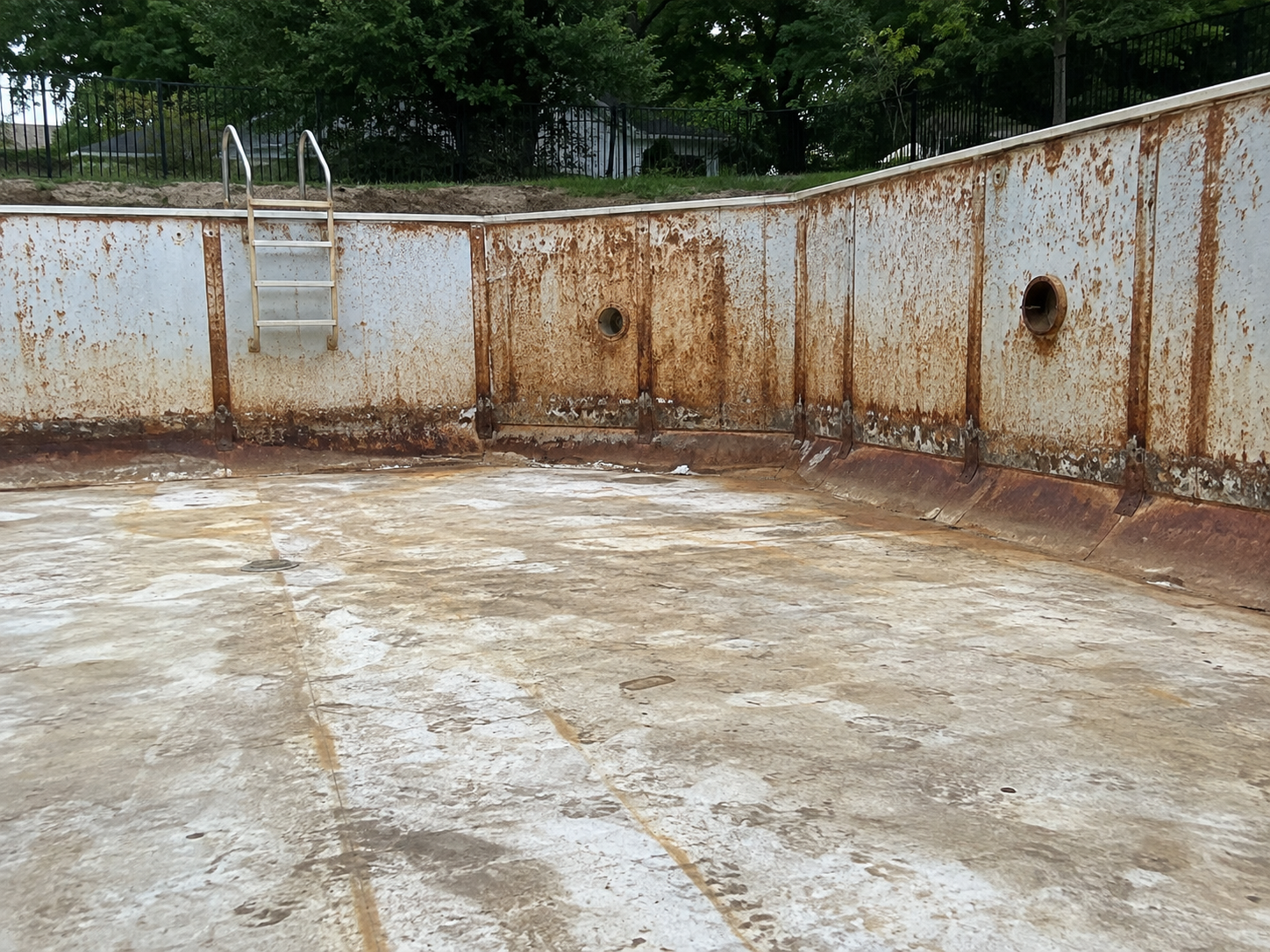 Empty, rusted swimming pool with a metal ladder, surrounded by a fence with trees in the background.