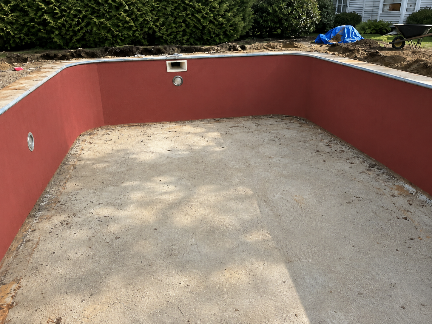 Empty swimming pool with red walls and a sandy bottom, under construction in a backyard.