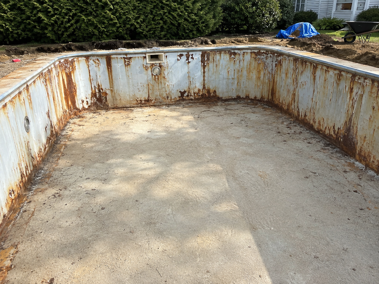 Old, rusty swimming pool with peeling paint and exposed metal, surrounded by a dirt area with construction materials and bushes in the background.