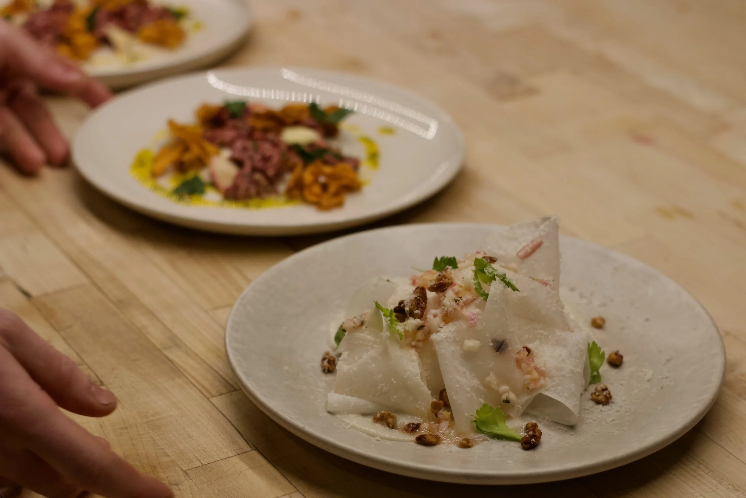 Close-up of a plate of food featuring a taco with chopped meat, cilantro, and other toppings, with additional plates of food in the background on a wooden table.