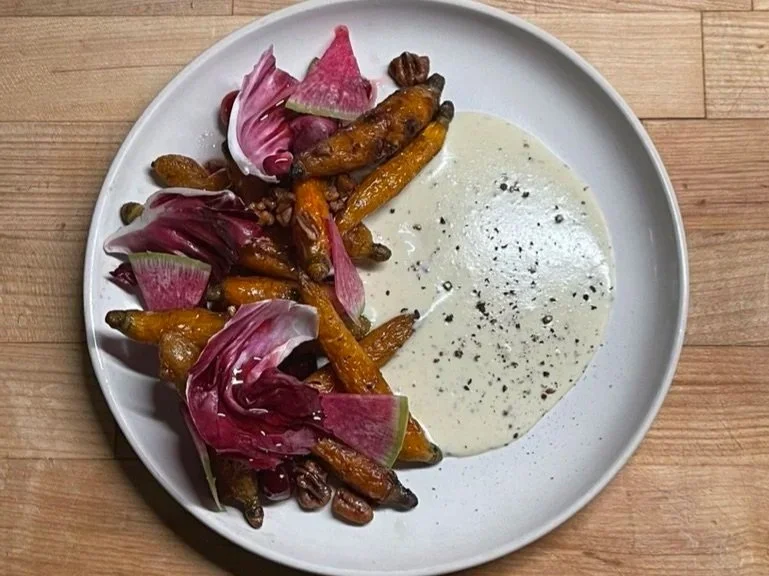 Plate with glazed carrots, radicchio leaves, and a side of creamy white dipping sauce with black pepper on a wooden table.