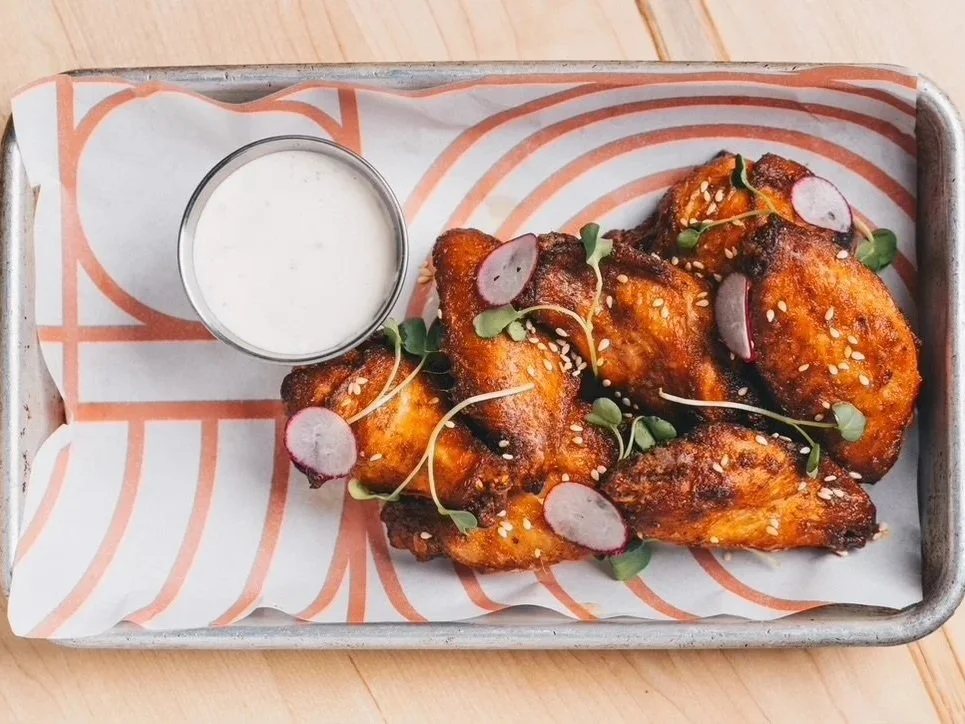 Baked chicken wings garnished with radish slices, microgreens, and sesame seeds, served with a side of ranch dipping sauce on a tray lined with striped paper.