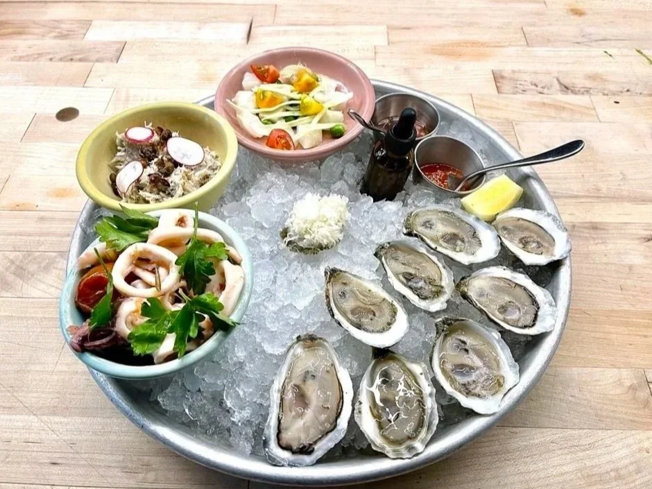 A metal tray filled with ice and fresh oysters on the half shell, surrounded by lemon wedges, hot sauce, and condiment bottles, with sides of seafood salads and vegetables in small bowls.