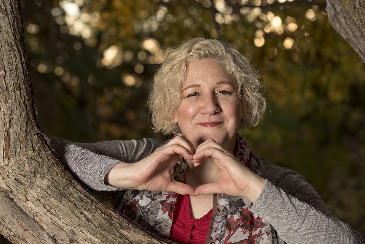A woman with curly blonde hair smiling and making a heart shape with her hands, leaning against a tree in an outdoor setting during fall.