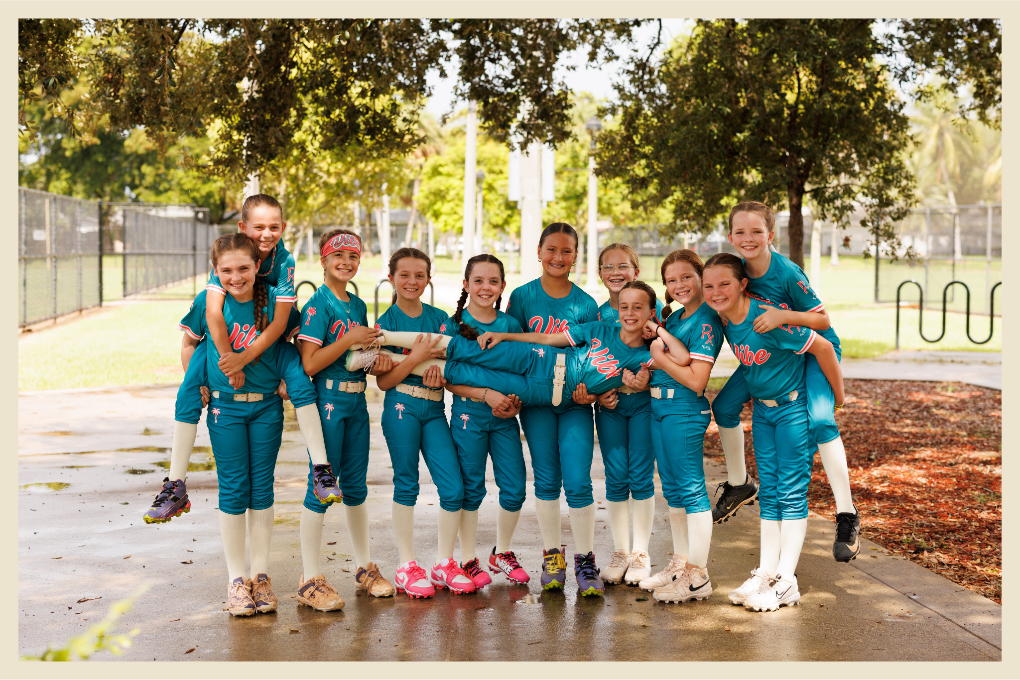 Eleven smiling youth softball players in teal “Vibe” uniforms pose together under a tree, with two teammates being held horizontally across the group.