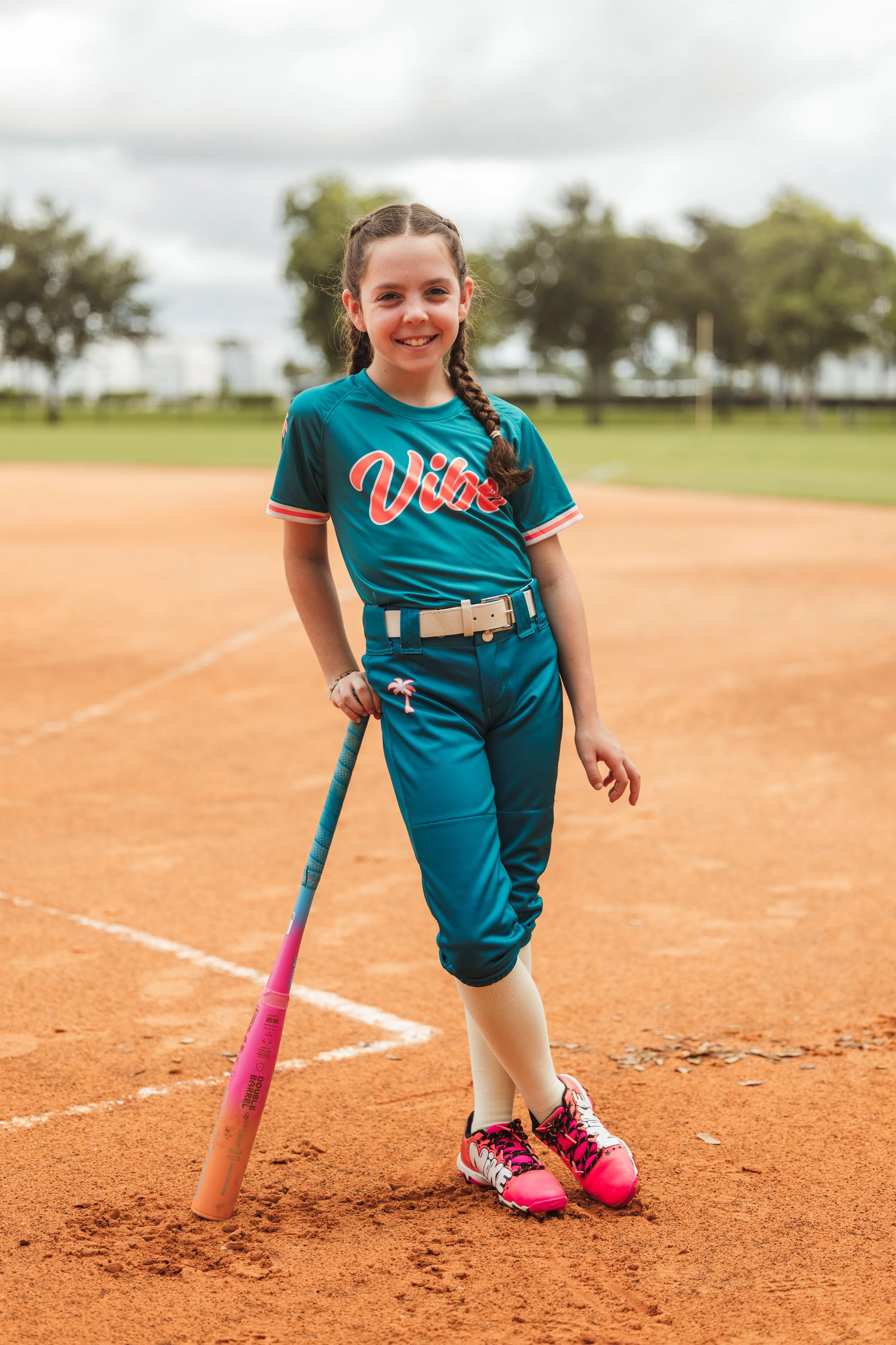Lily, Vibe softball player number 8, smiles while posing with a colorful bat on the infield.