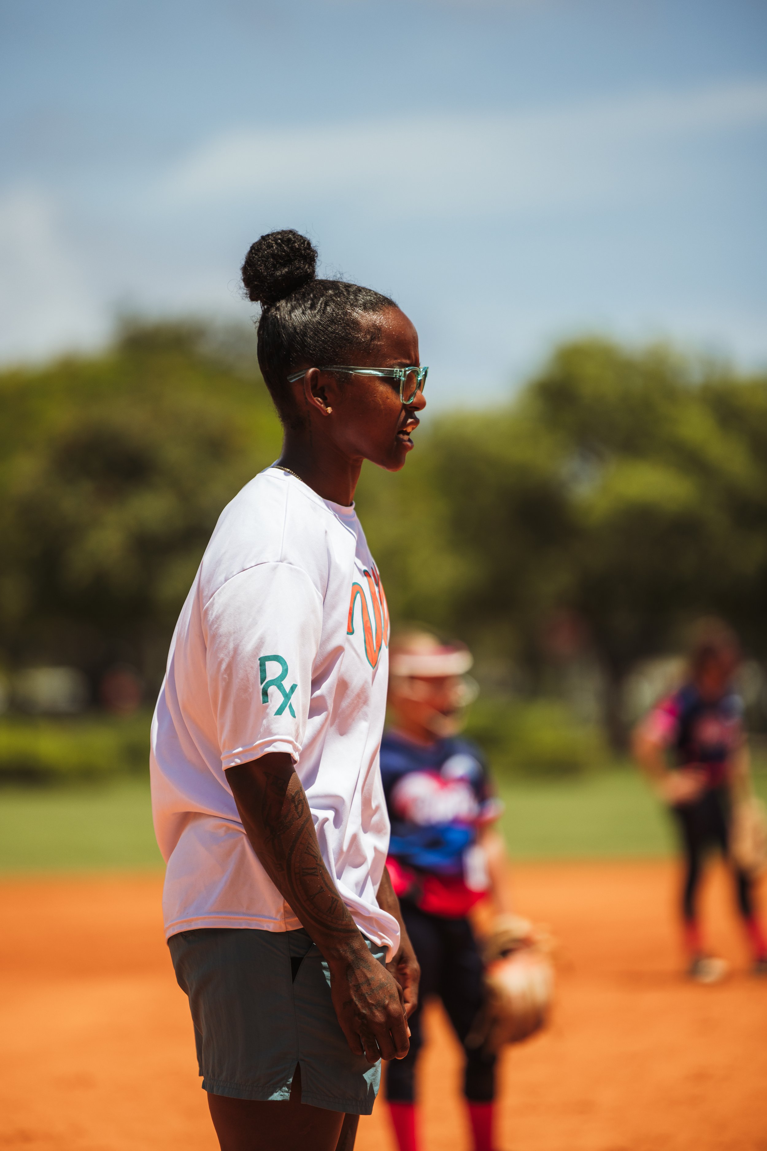 A woman with glasses and a bun hairstyle stands on a baseball field during daytime, surrounded by children in sports uniforms, with trees in the background.