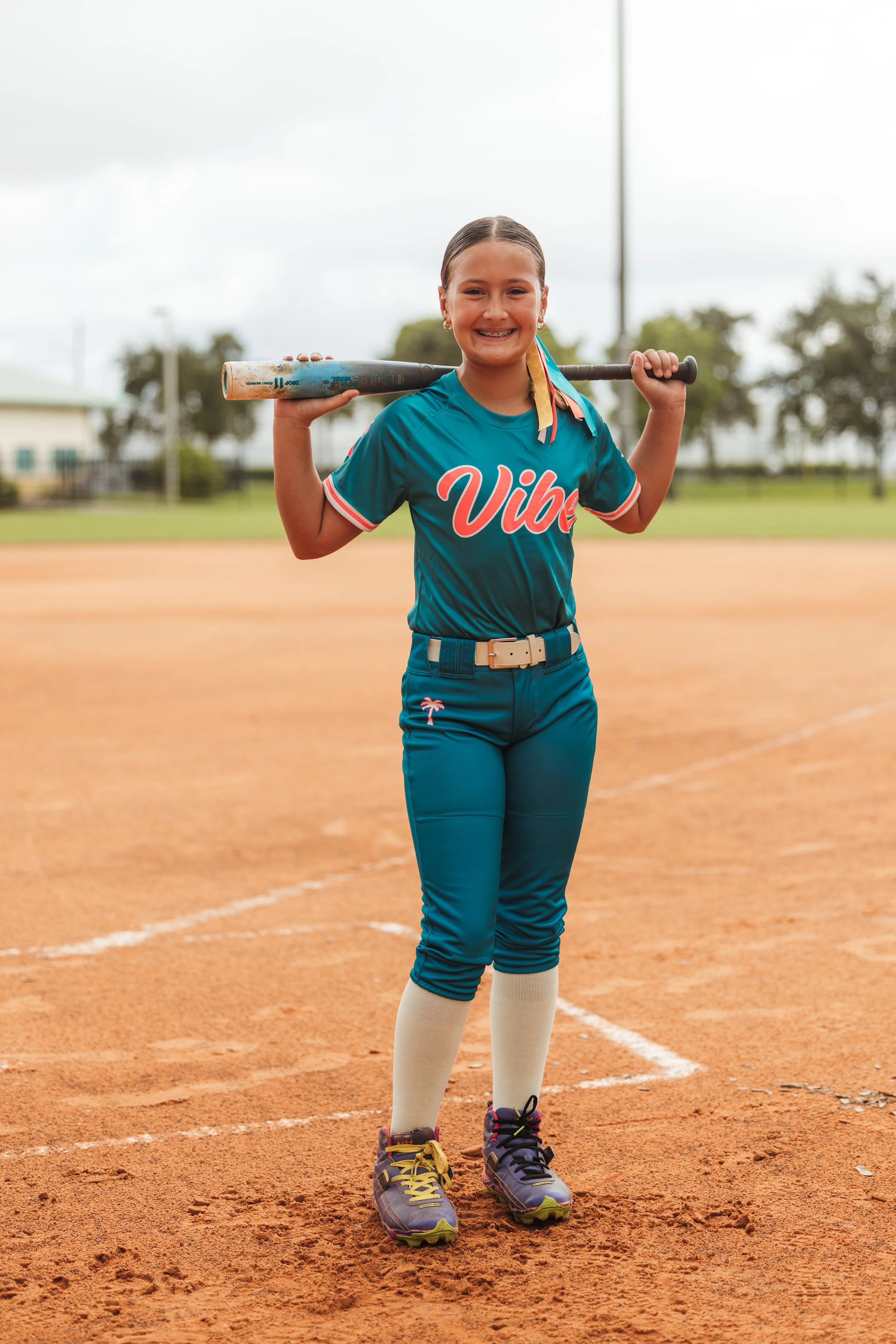 Gisele, Vibe softball player number 6, smiles on the infield with a bat resting across her shoulders.
