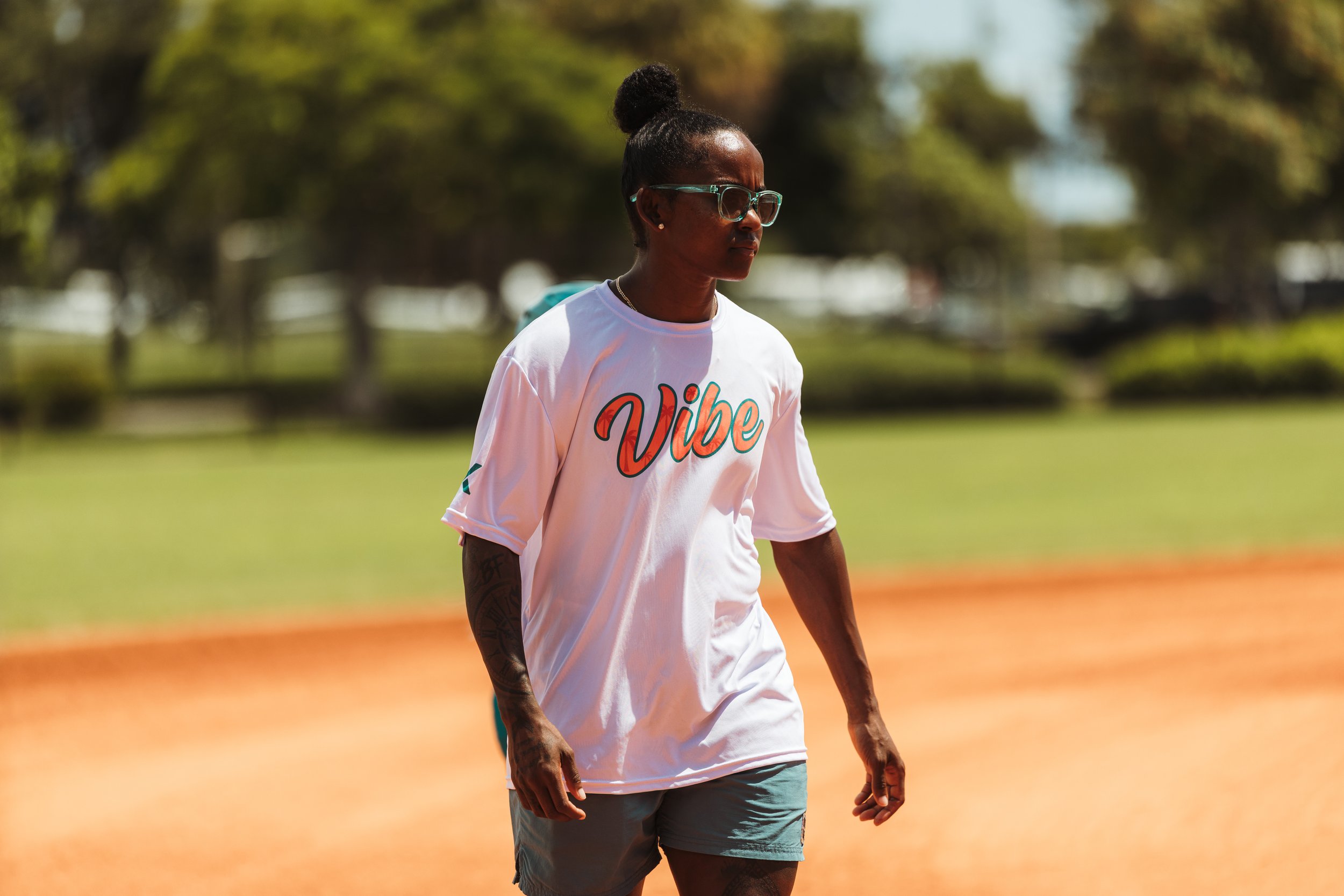 Vibe softball coach wearing sunglasses and a white team shirt walks across the infield.