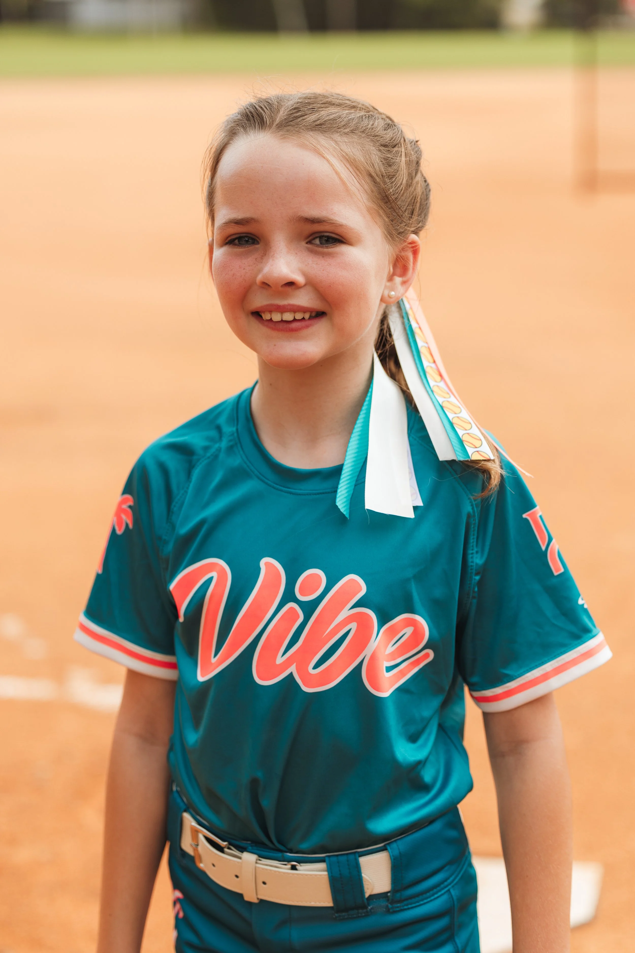 Ava, Vibe softball player number 13, smiles for a portrait in her teal uniform on the field.