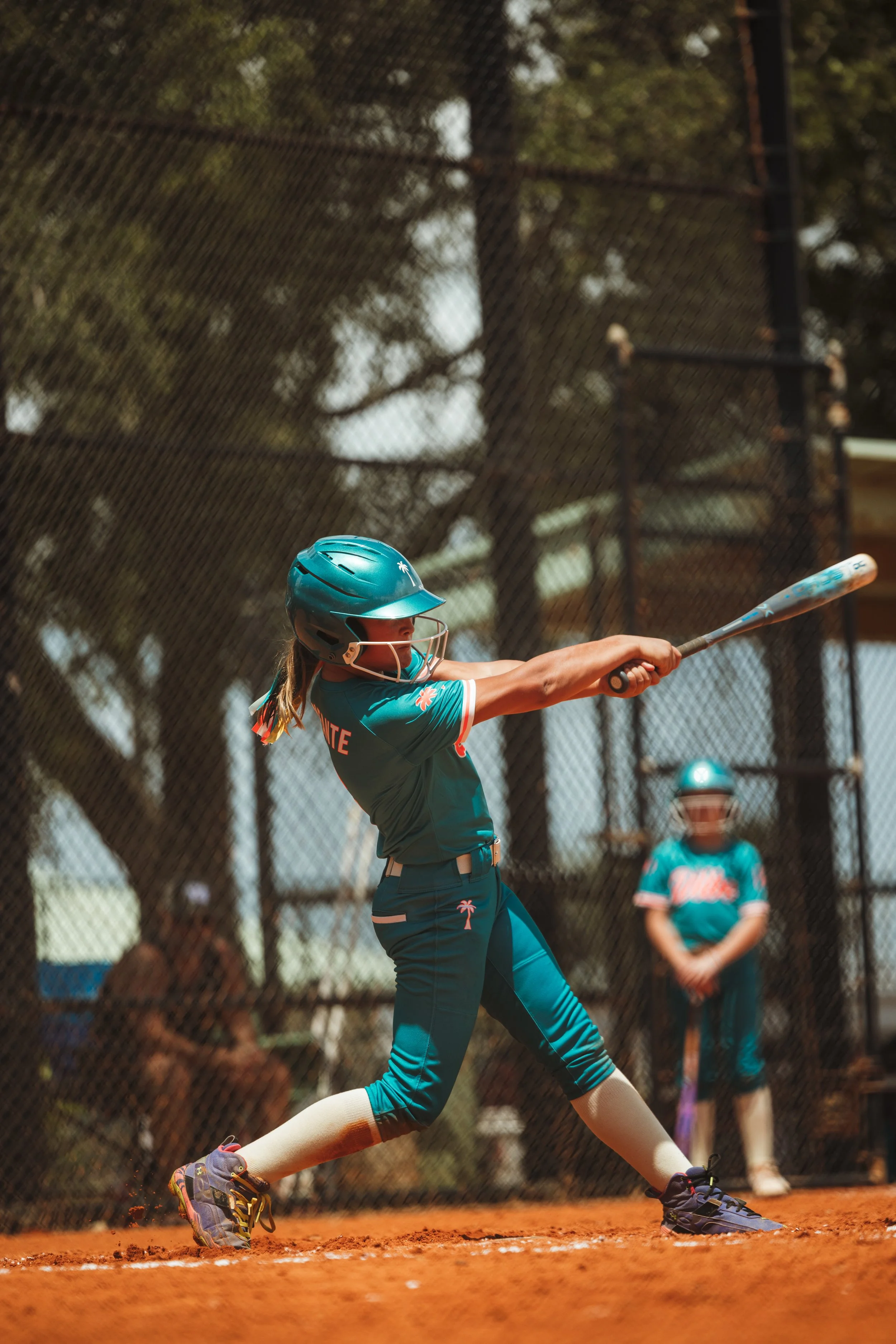 Youth Vibe softball player in a teal uniform and helmet swings a bat at home plate.