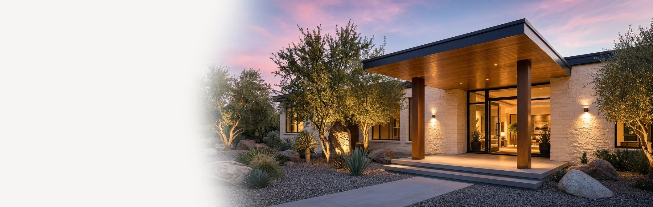 Modern house with a covered entrance, surrounded by desert landscape with trees and rocks, during dusk.