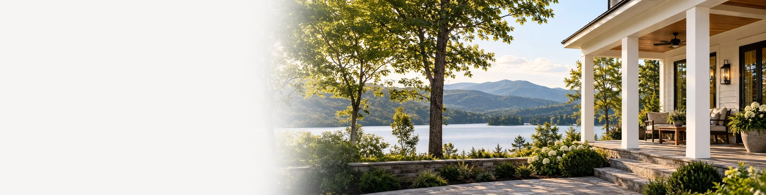 Porch of a house with outdoor seating, overlooking a lake and mountain landscape with trees and blue sky.