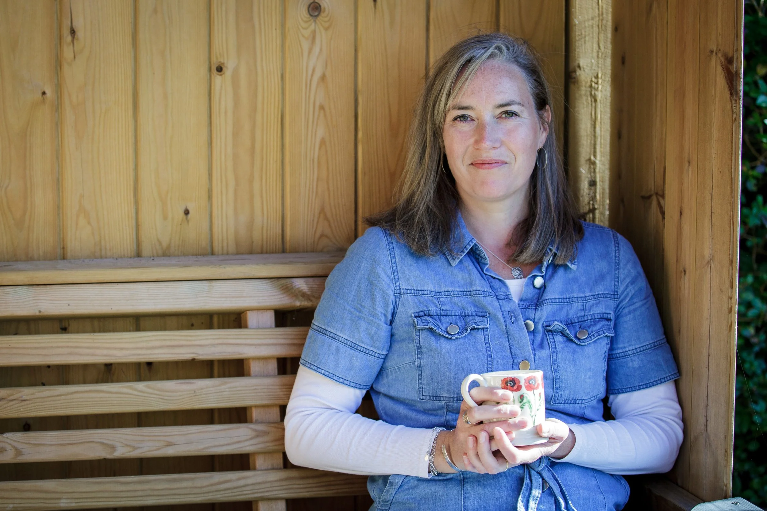 Woman with long brown hair, wearing a denim dress and holding a floral mug, sitting in a wooden summerhouse outdoors.