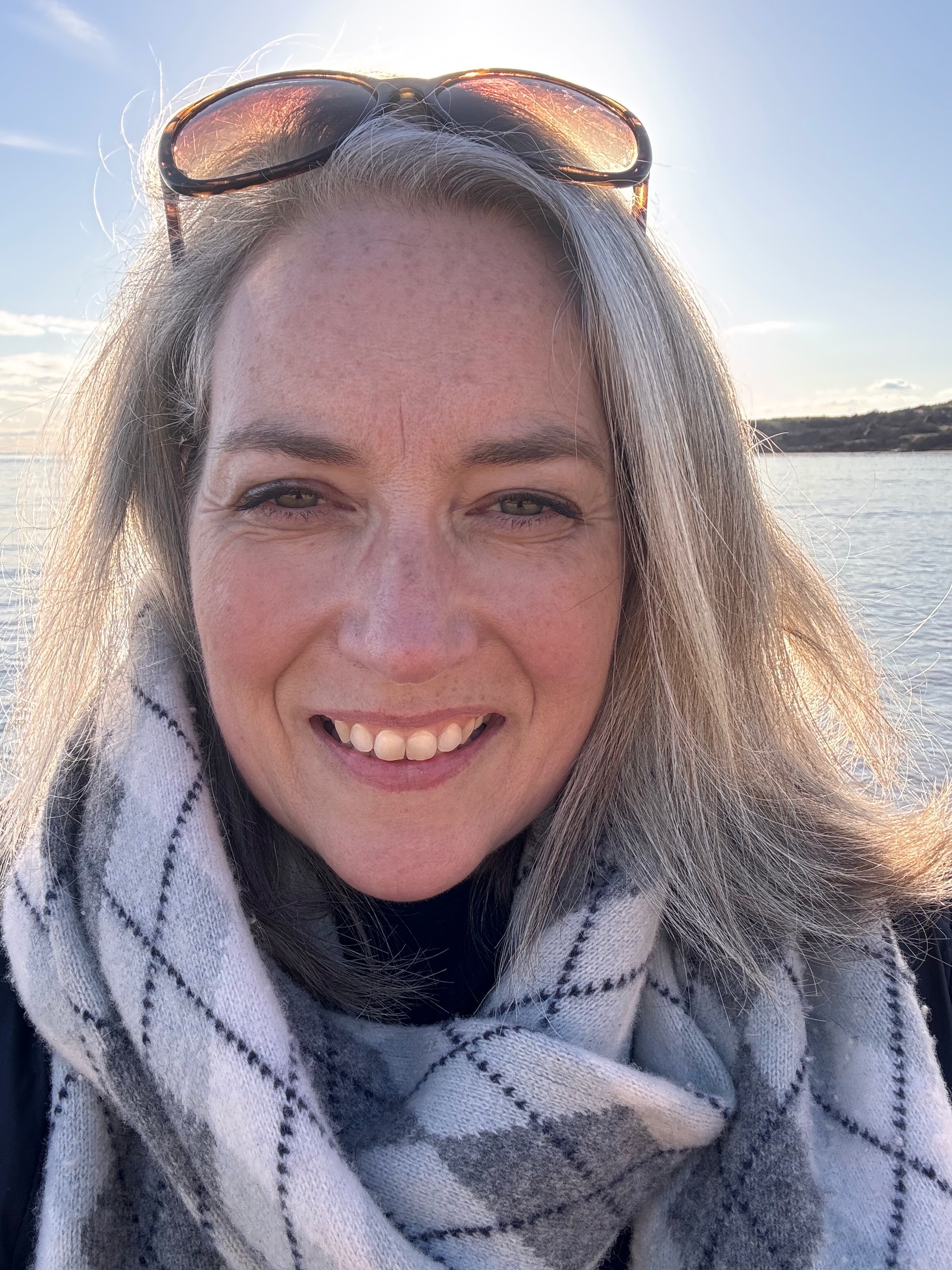 Close-up of a smiling woman with gray hair, wearing sunglasses on her head and a cream-colored scarf with black stripes, outdoors by a body of water with a landscape and cloudy sky.