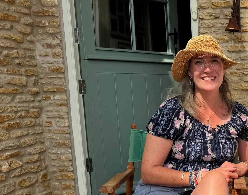 A woman with gray hair wearing a straw hat and floral blouse, smiling while sitting outside near a stone wall and a blue door.