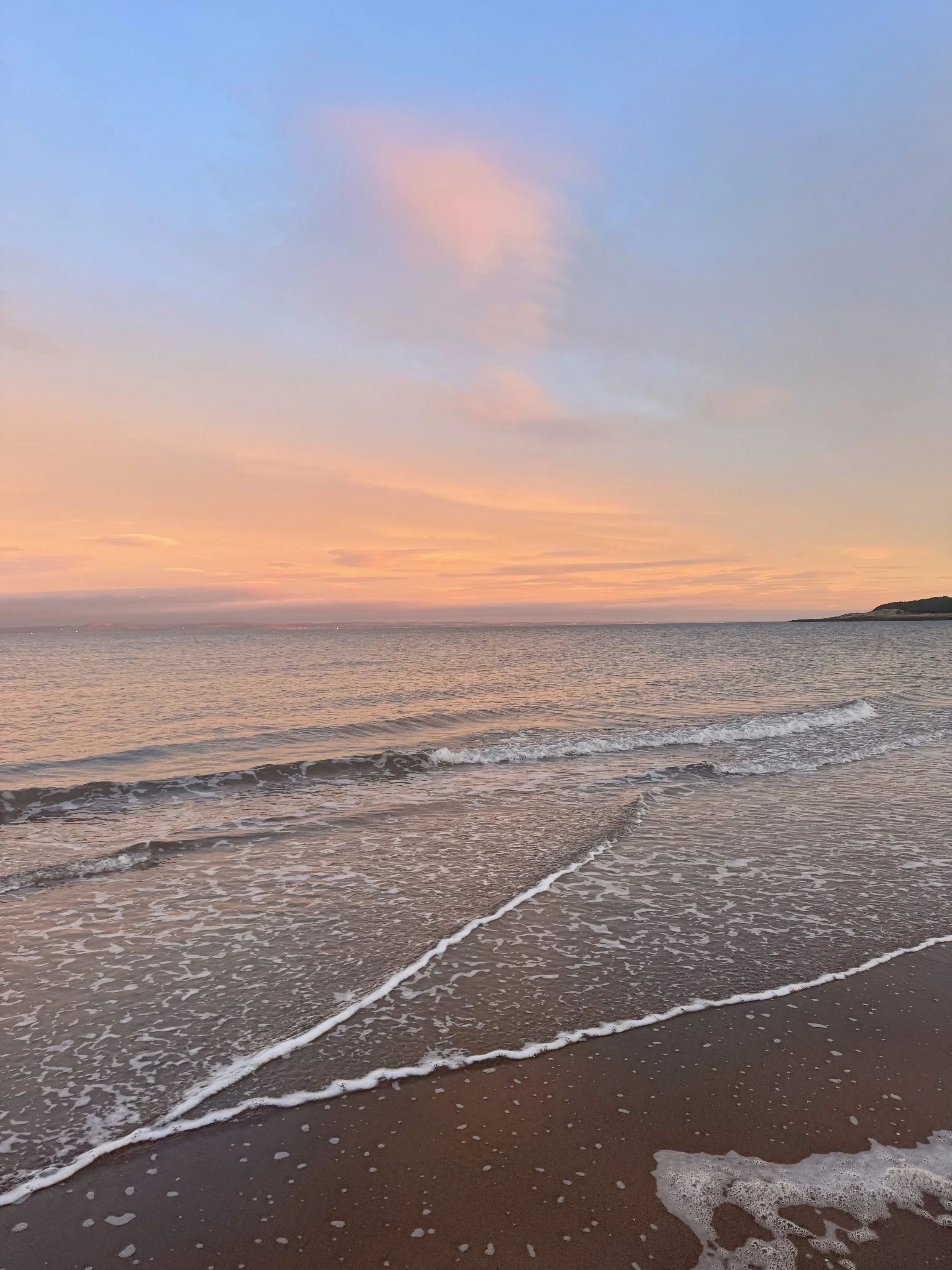 Sunset over the ocean with pastel-colored sky, gentle waves on the sandy beach, and a distant landmass on the horizon.