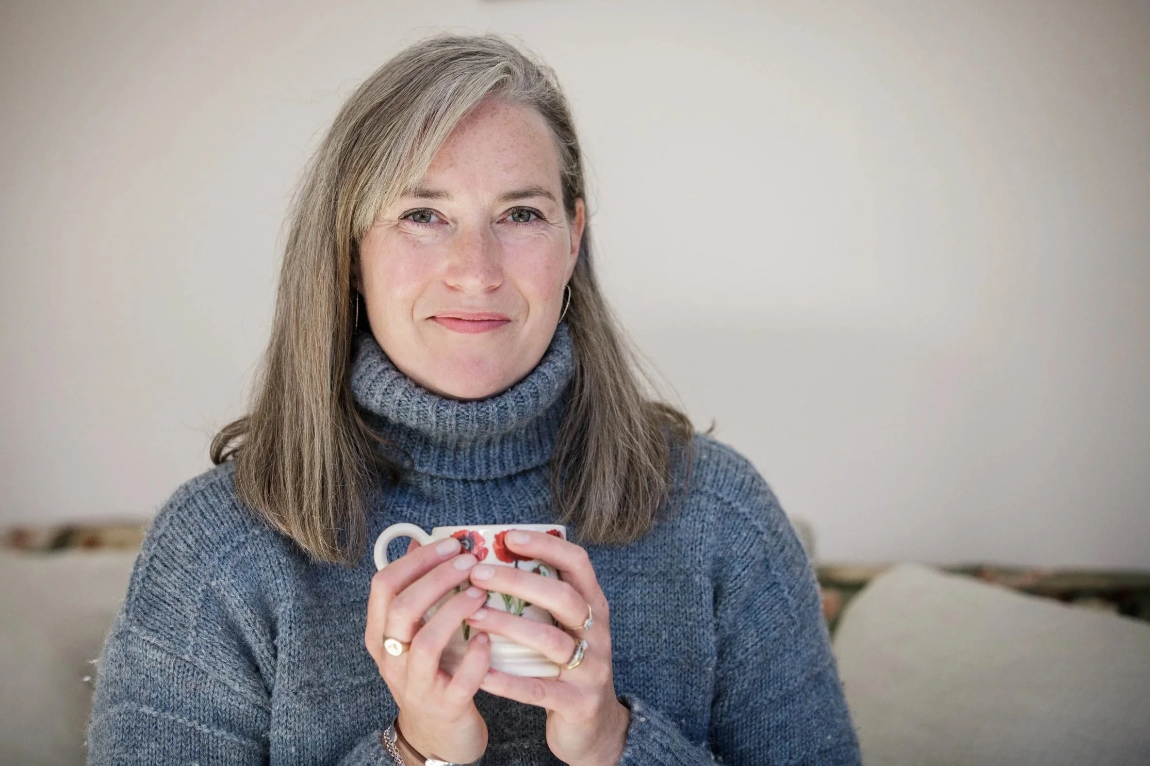 A woman with long light brown hair and a gray turtleneck sweater, holding a floral mug with both hands, smiling gently, in a cozy indoor setting.
